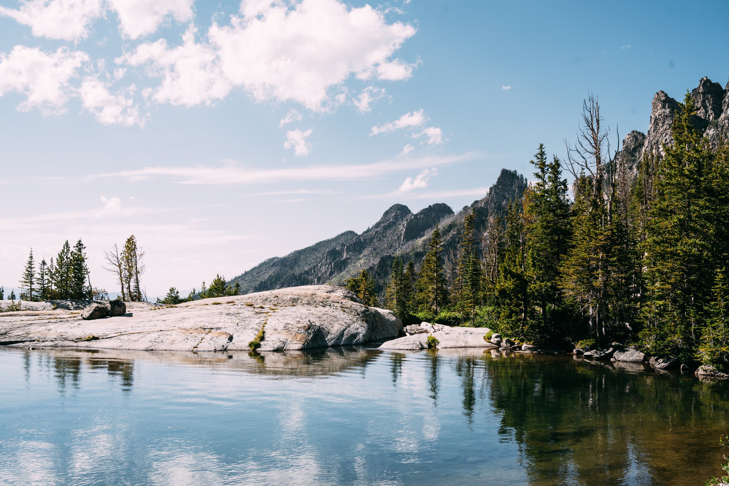 A serene mountain lake scene with calm water reflecting the trees and mountains in the background, under a partly cloudy sky.