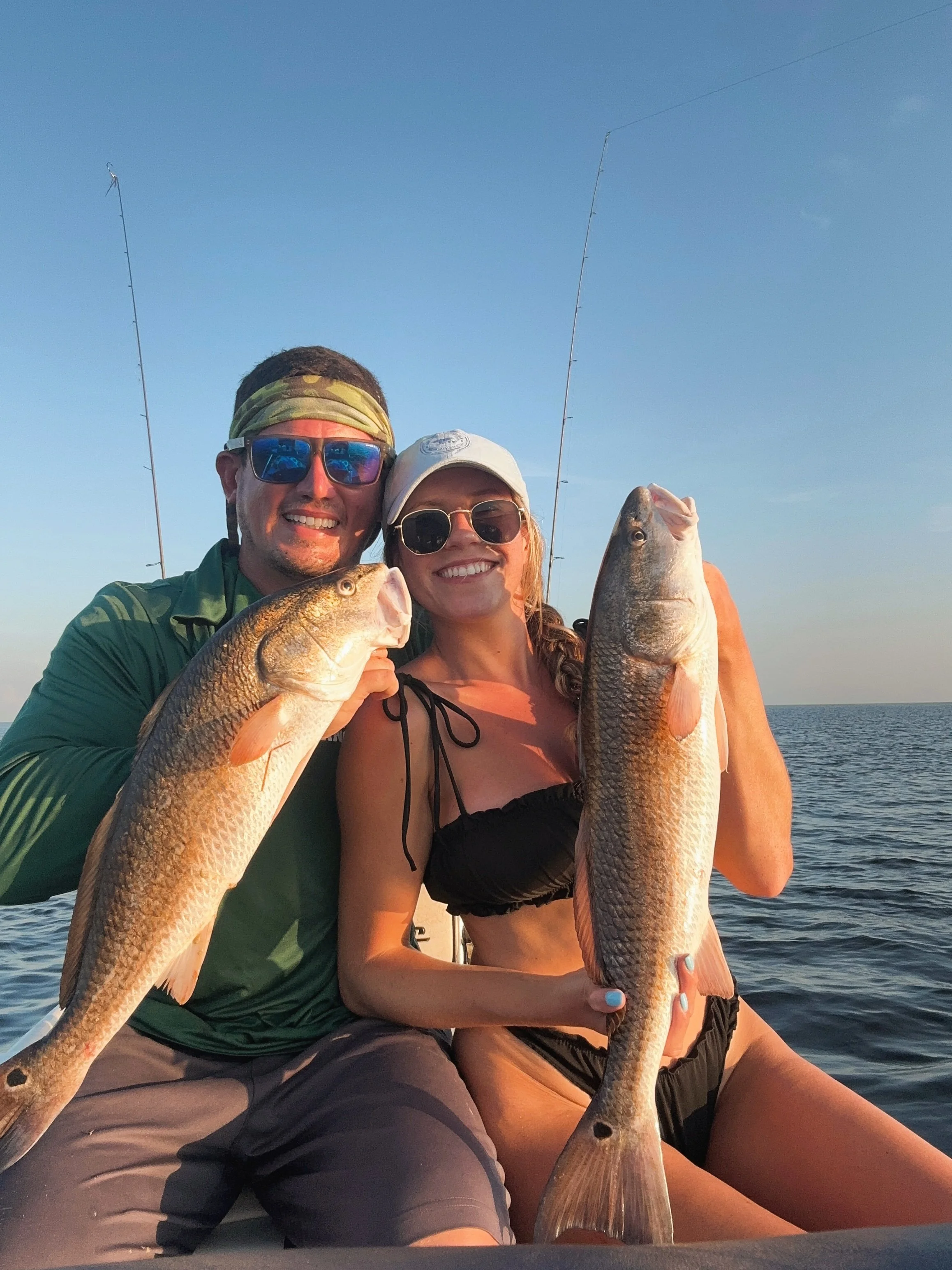 Two people on a boat holding up two large fish on a sunny day with the ocean in the background.