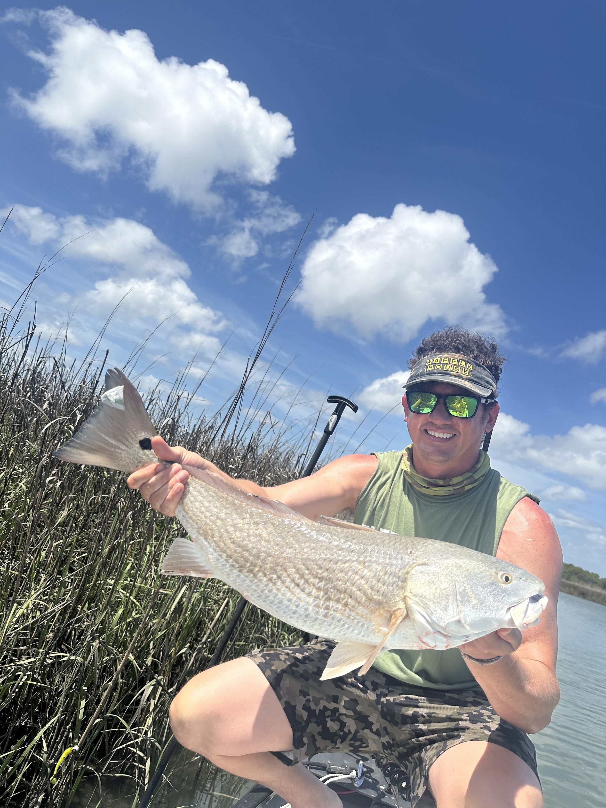 A man wearing sunglasses, a visor, and sleeveless shirt, kneeling outdoors near water and tall grass, holding a large fish with silvery scales and a prominent dorsal fin.