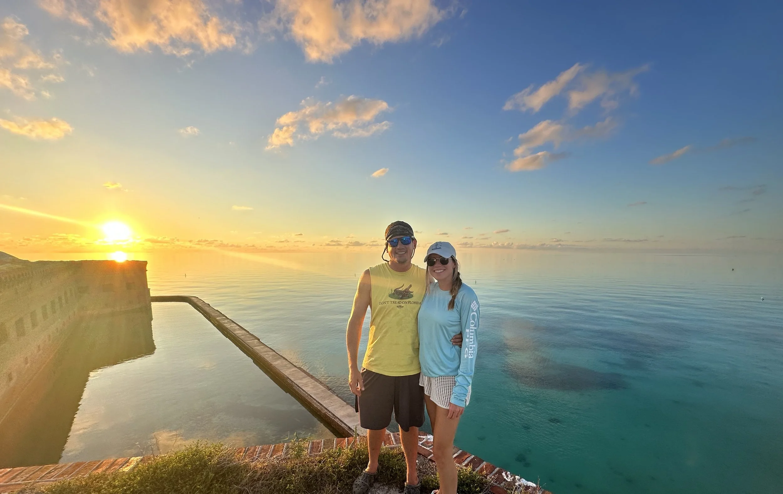 A man and woman standing together near a body of water during sunset, smiling. The man is wearing a yellow tank top and dark shorts, while the woman is in a light blue long sleeve shirt and shorts. Both are wearing sunglasses and hats.