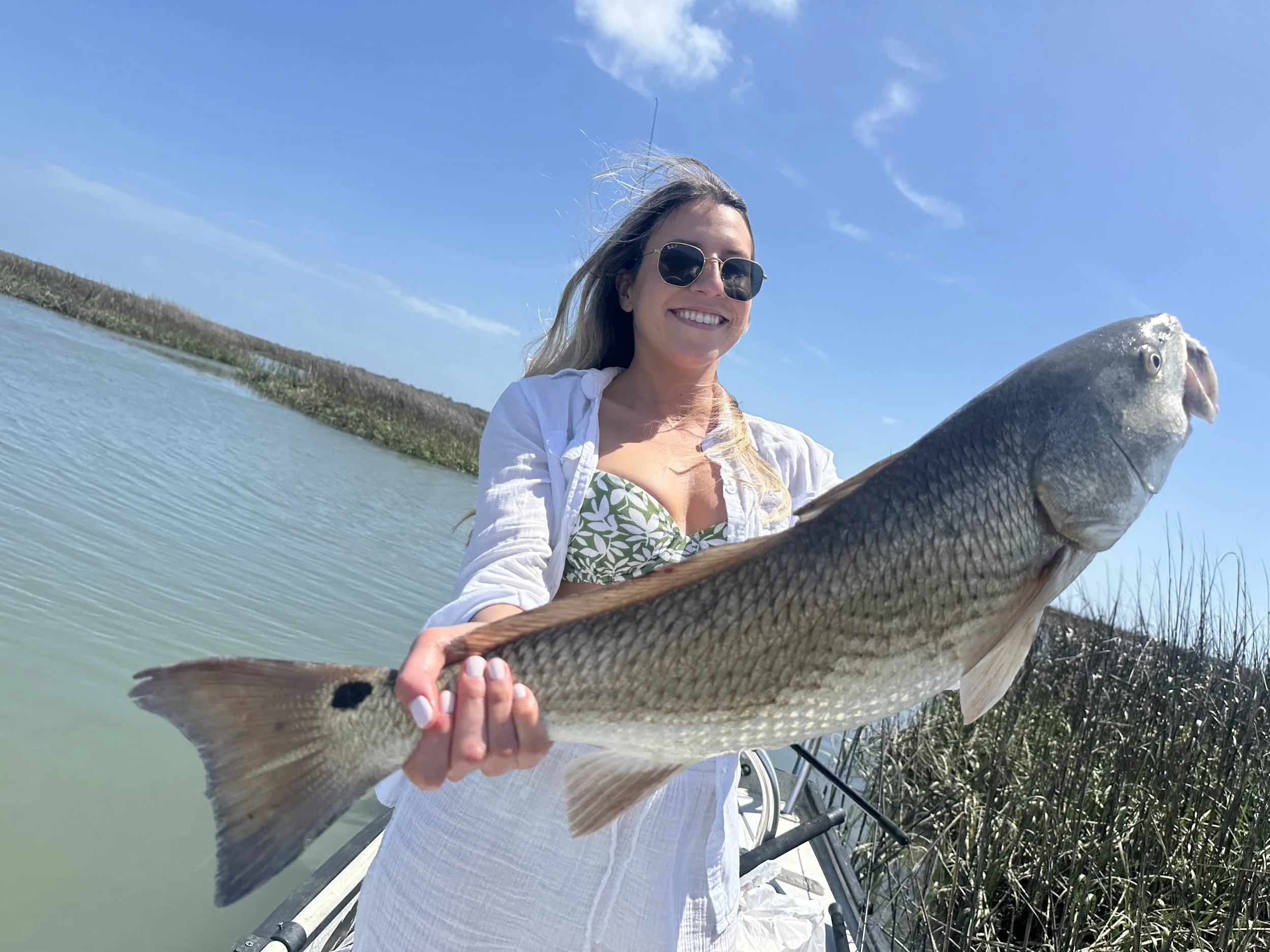 Woman wearing sunglasses holding a large redfish on a boat in a marsh area under a partly cloudy sky.