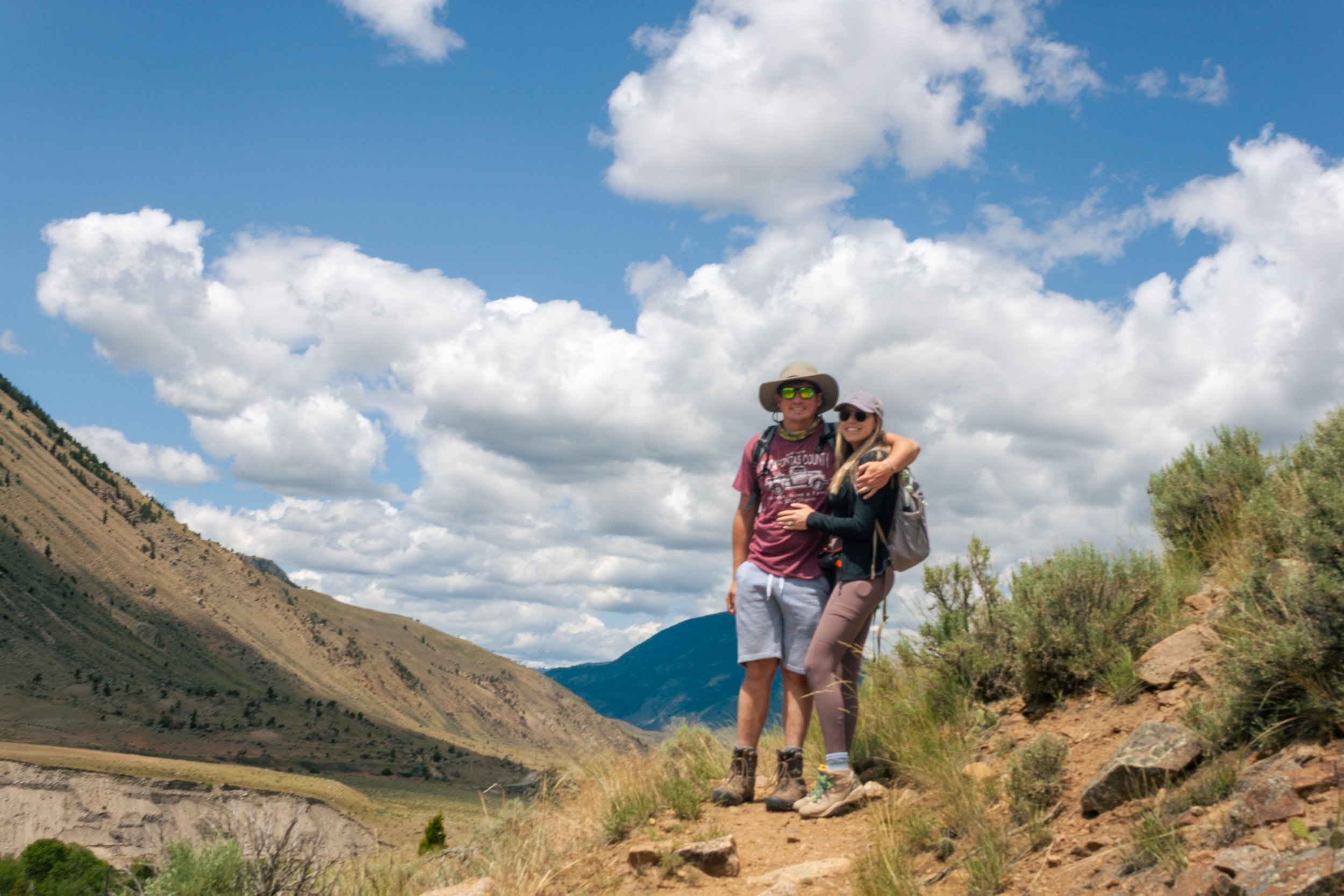 A man and woman standing together on a mountain trail, smiling and hugging, with mountains and a partly cloudy sky in the background.