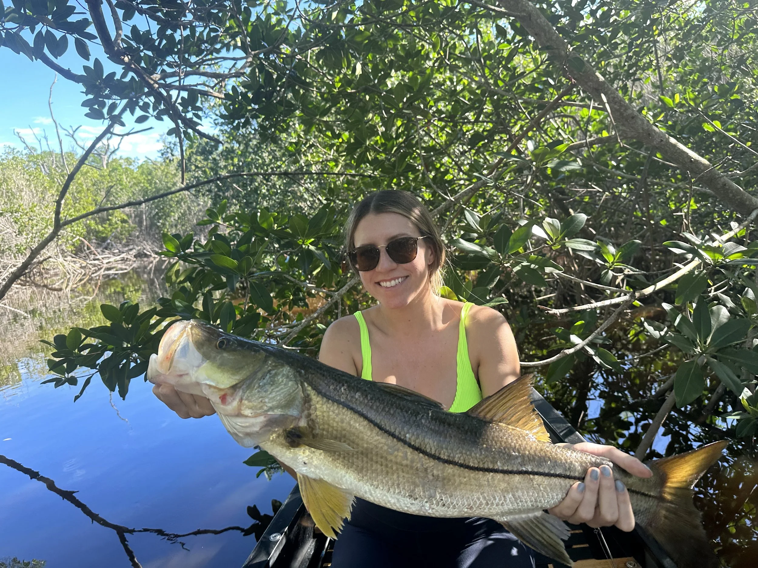 A woman wearing sunglasses and a neon green tank top smiling while holding a large fish with a waterway and dense greenery in the background.