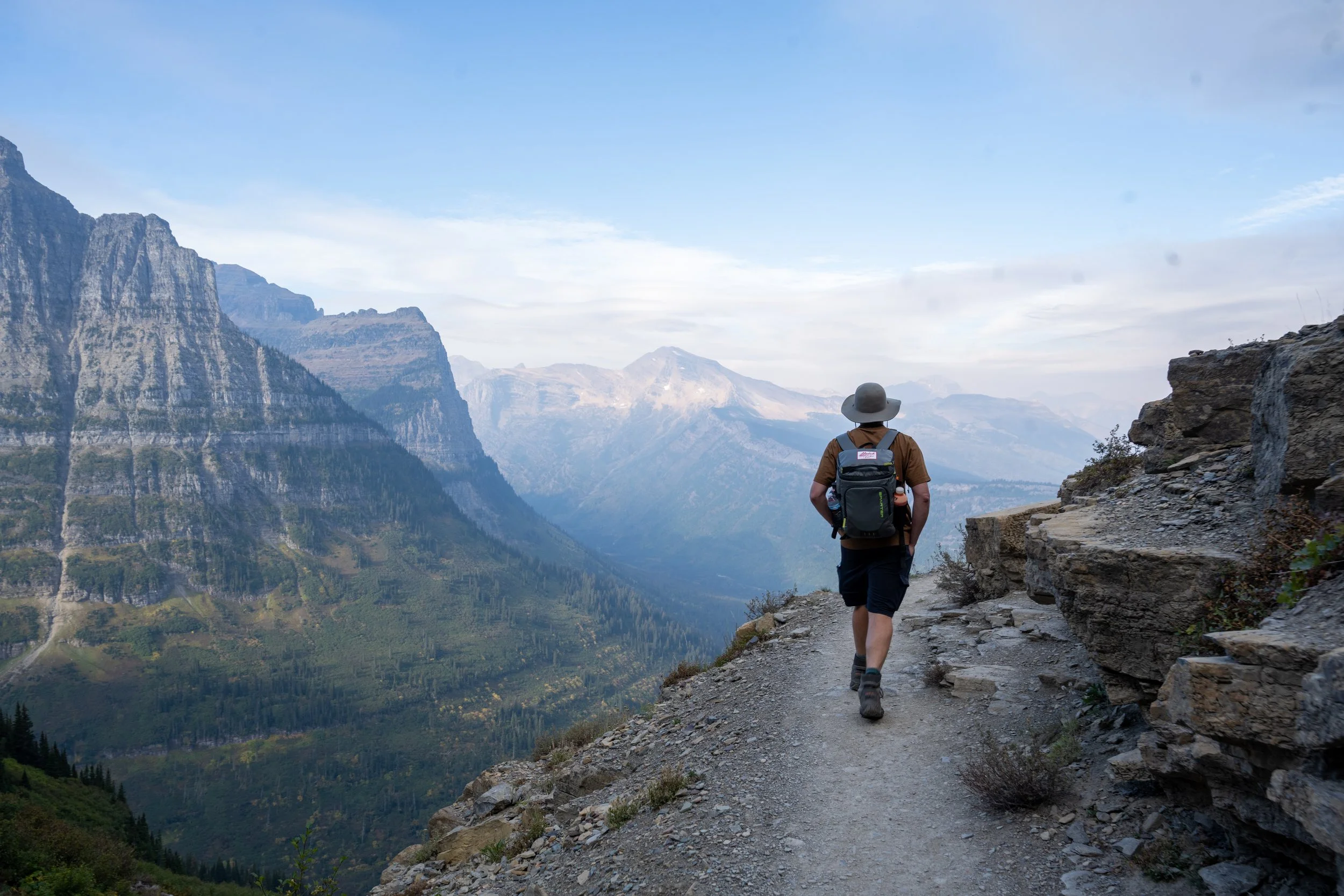 Person hiking on a mountain trail with scenic mountain ranges and a cloudy sky in the background.