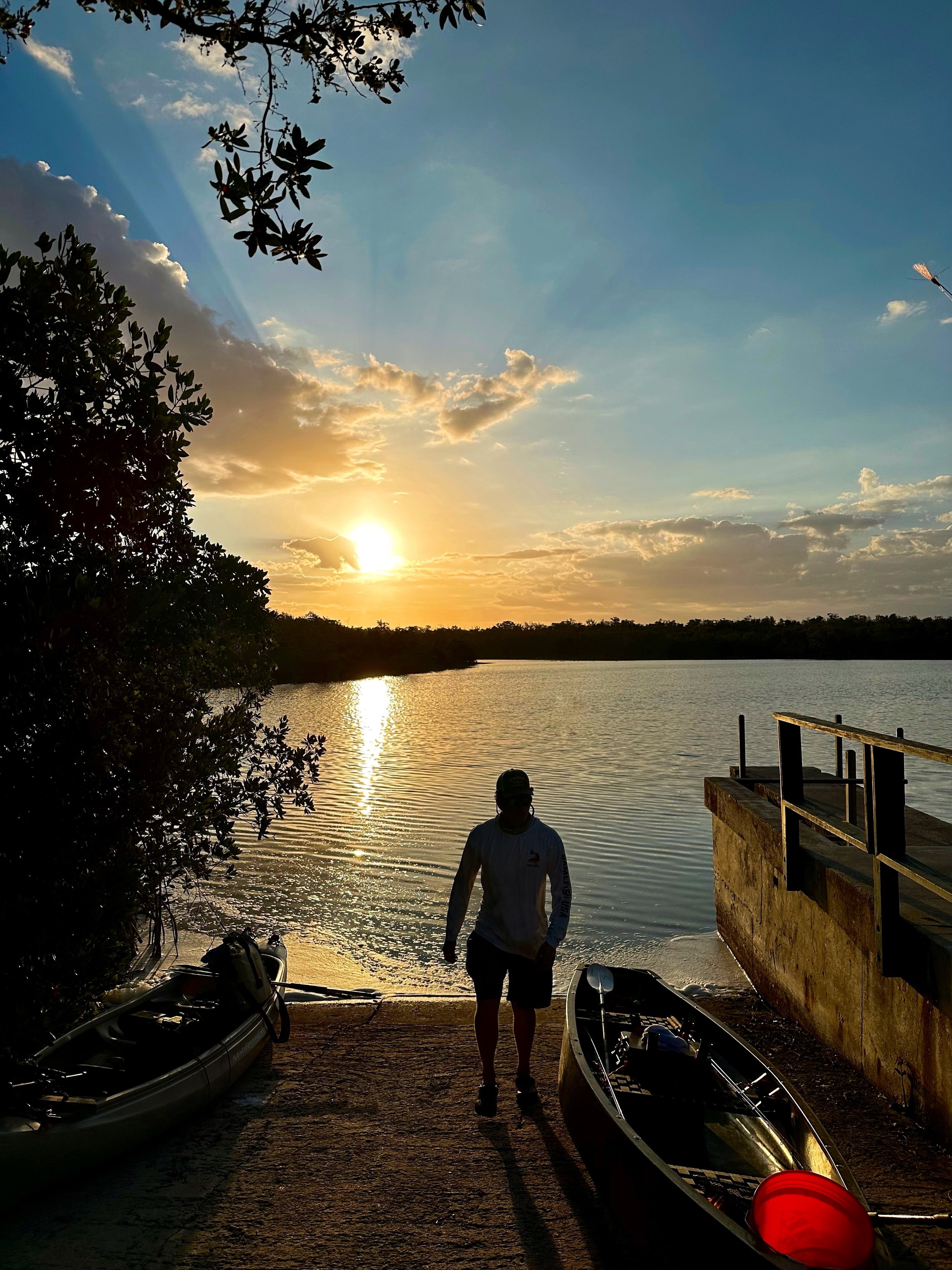 Silhouette of a person standing on a boat ramp at sunset with two kayaks nearby, overlooking a calm river with trees in the background and clouds in the sky.