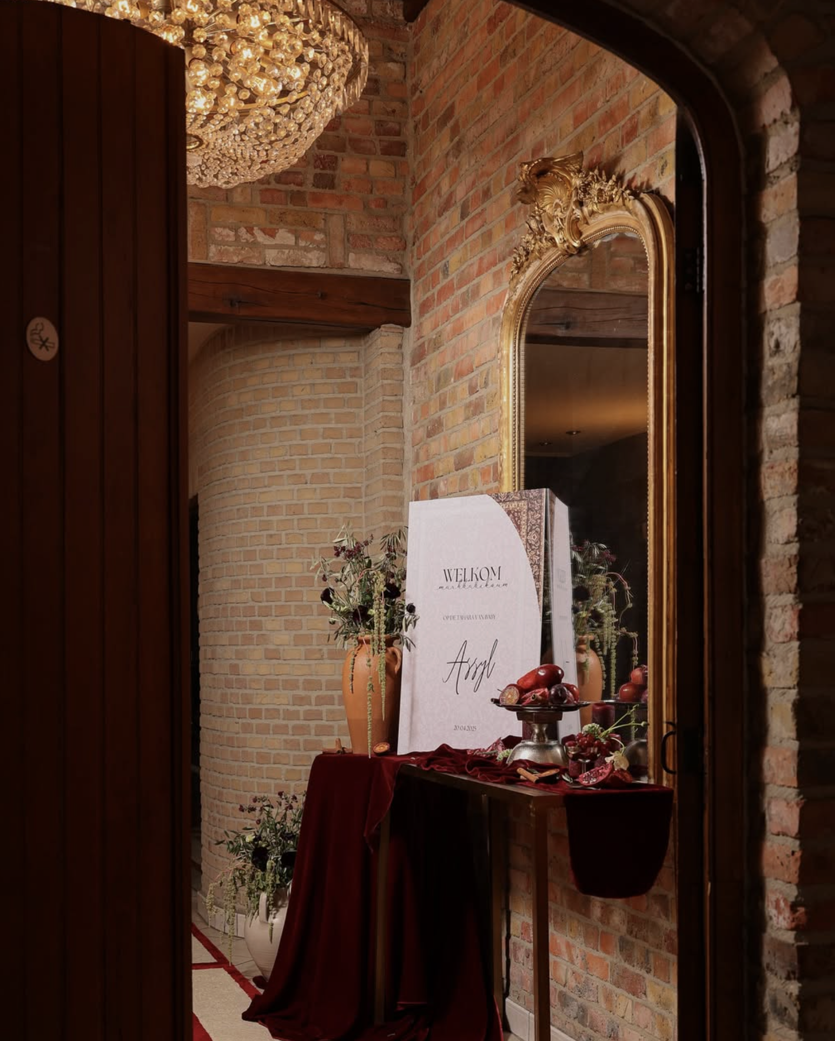 Een decoratief interieur met een grote spiegel in een gouden lijst, bloemen in vazen, een welkomstbord met tekst, en een tafel bedekt met een rood doek en fruit, in een kamer met bakstenen muren en een kristallen chandelier.