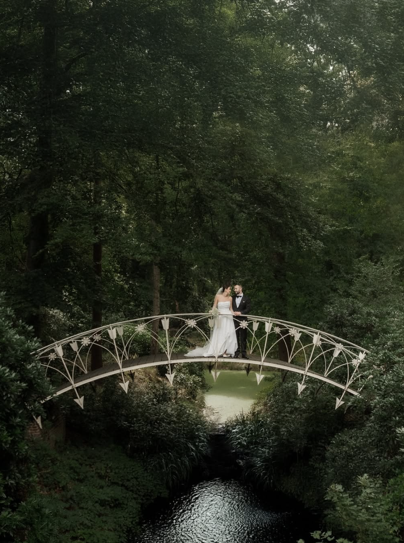 Een net getrouwde stel op een witte brug over een kleine rivier in een bosrijke omgeving.