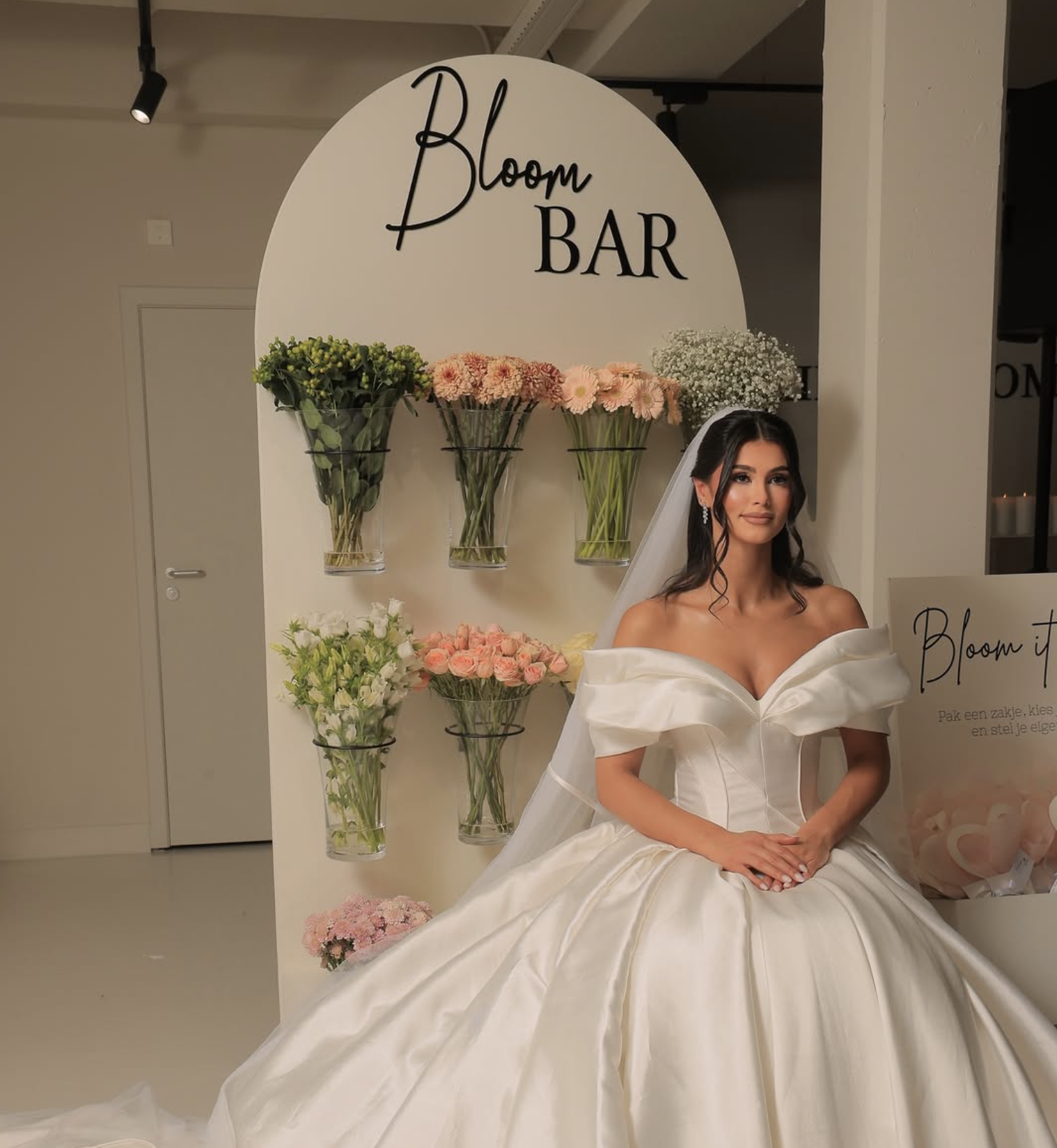 Een vrouw in een witte bruiloftsjurk zit voor een bloemwinkel met een bord dat 'Bloom BAR' zegt, omringd door vazen met bloemen.