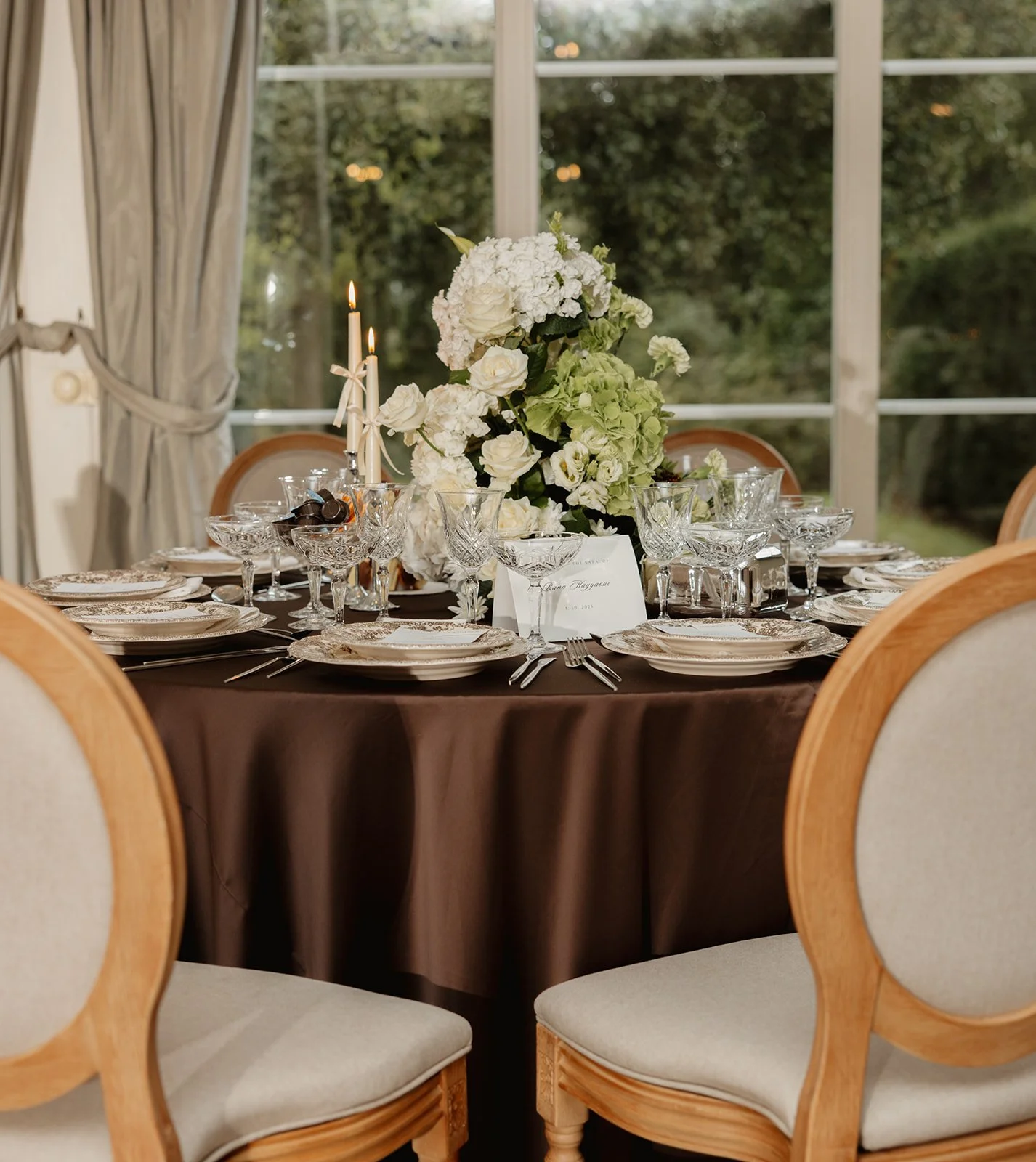 Een ronde eettafel bedekt met een donkerbruine tafelkleed, versierd met een grote bloemarrangement van witte en groene bloemen in het midden, omgeven door kristallen glaswerk, borden, bestek en een kaartje, met een venster en groene bomen op de achte