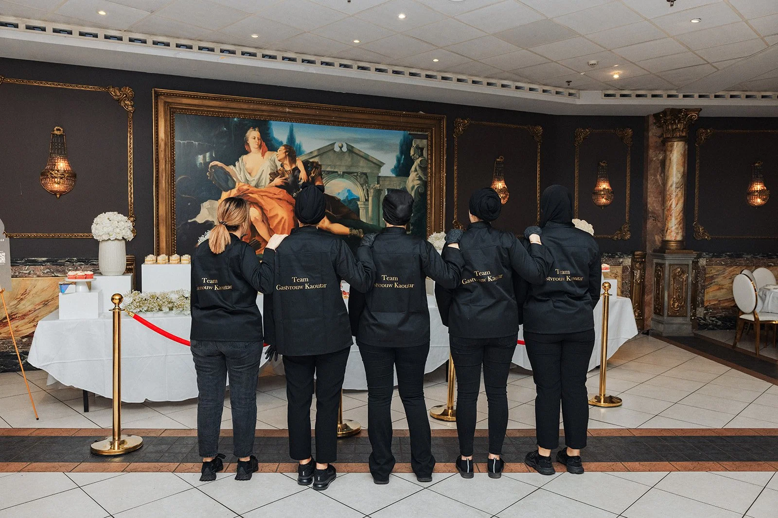 Vijf vrouwen in zwarte uniformen met kapjes kijken naar een grote schilderij in een elegante ruimte, met witte tafels en decoratieve wandlampen.