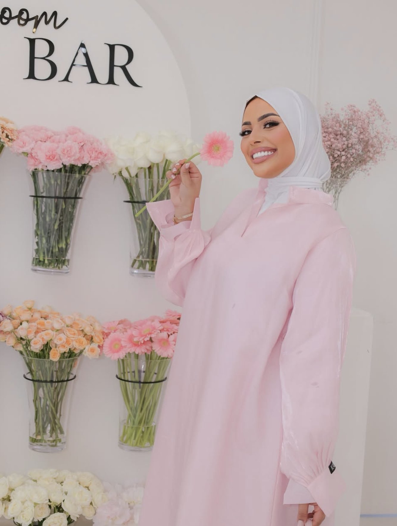 Vrouw in een witte hoofddoek en lichtroze jurk poseert bij een bloemwand, houdt een pinke bloem vast en glimlacht.