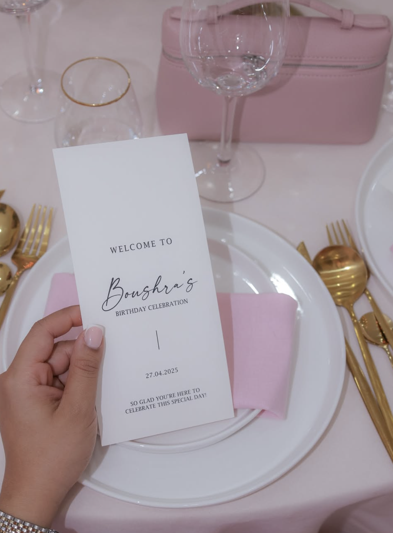 Een pink en wit gedekte feesttafel met een uitnodiging voor Boushra's verjaardag, glazen, goudkleurige bestekken, een roze tas, en een bord op een roze tafellaken.