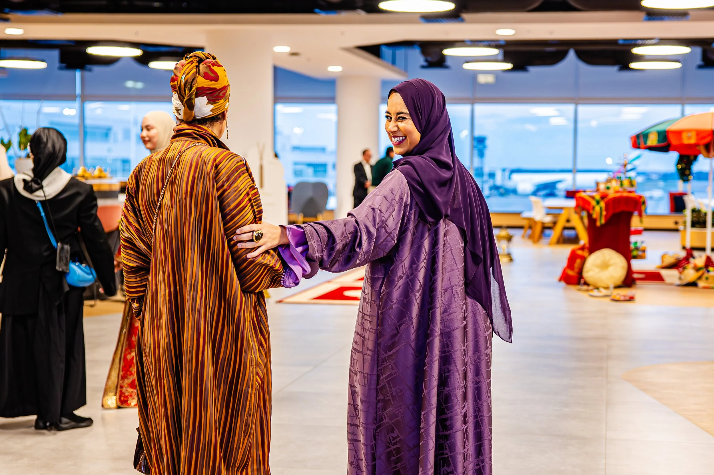 Twee vrouwen in kleurrijke traditionele hoofddoeken en kleding die elkaar bij de arm vasthouden en lachen in een luchthavenhal met grote ramen en uitzicht op vliegtuigen.