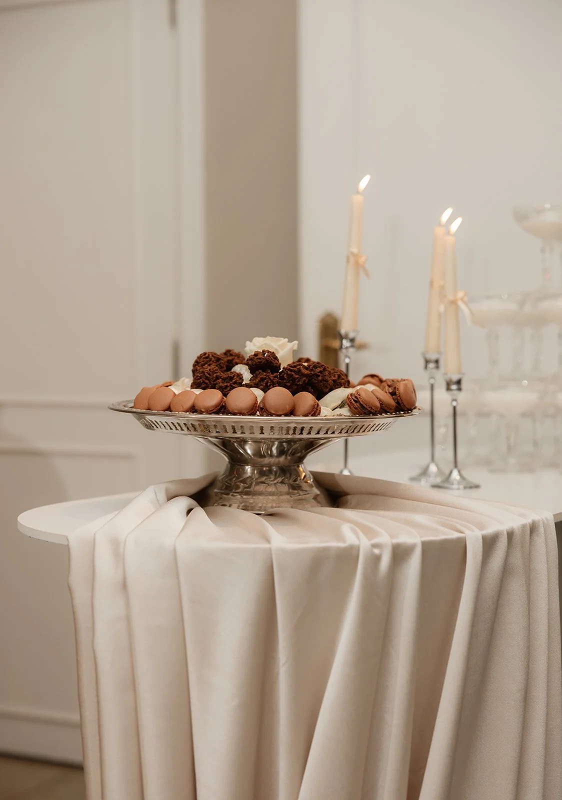 Elegante tafel met een zilveren schaal vol chocolade truffels en macarons, versierd met witte bloemen, op een avondfeest met kaarslicht en glazen kandelaren op de achtergrond.