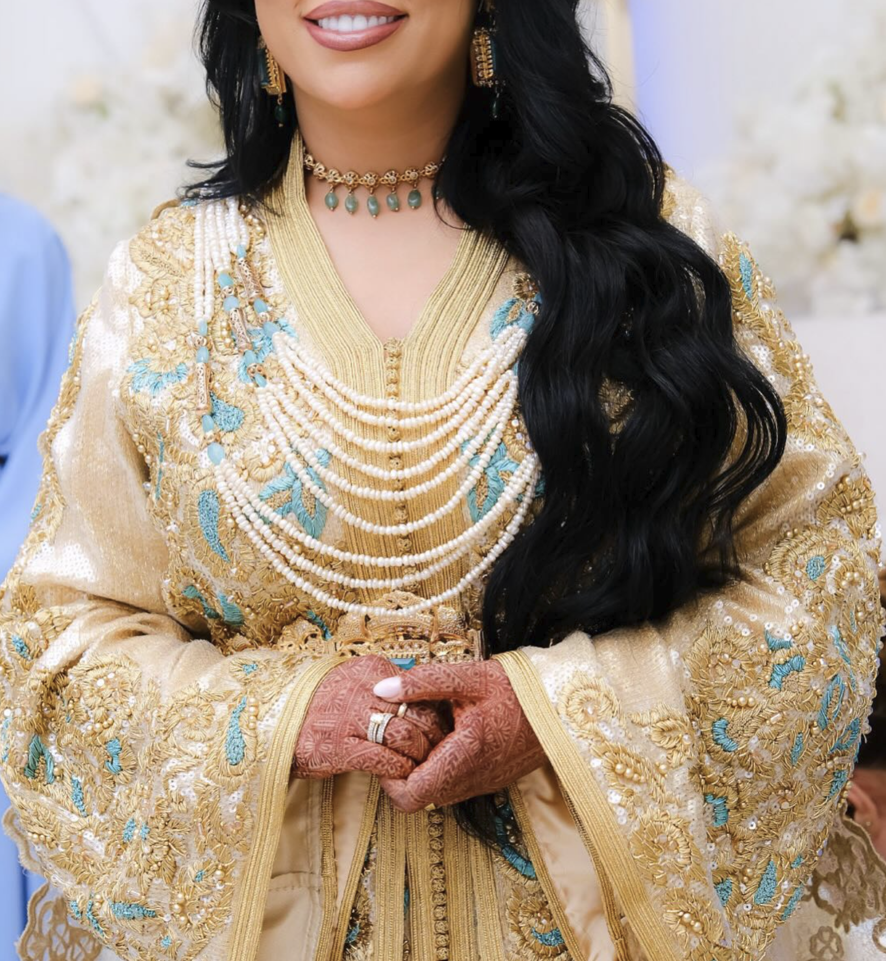Een vrouw in een gouden en blauwe embroidered jurk, gedragen met meerdere parelkettingen, haar handen met henna en sieraden.