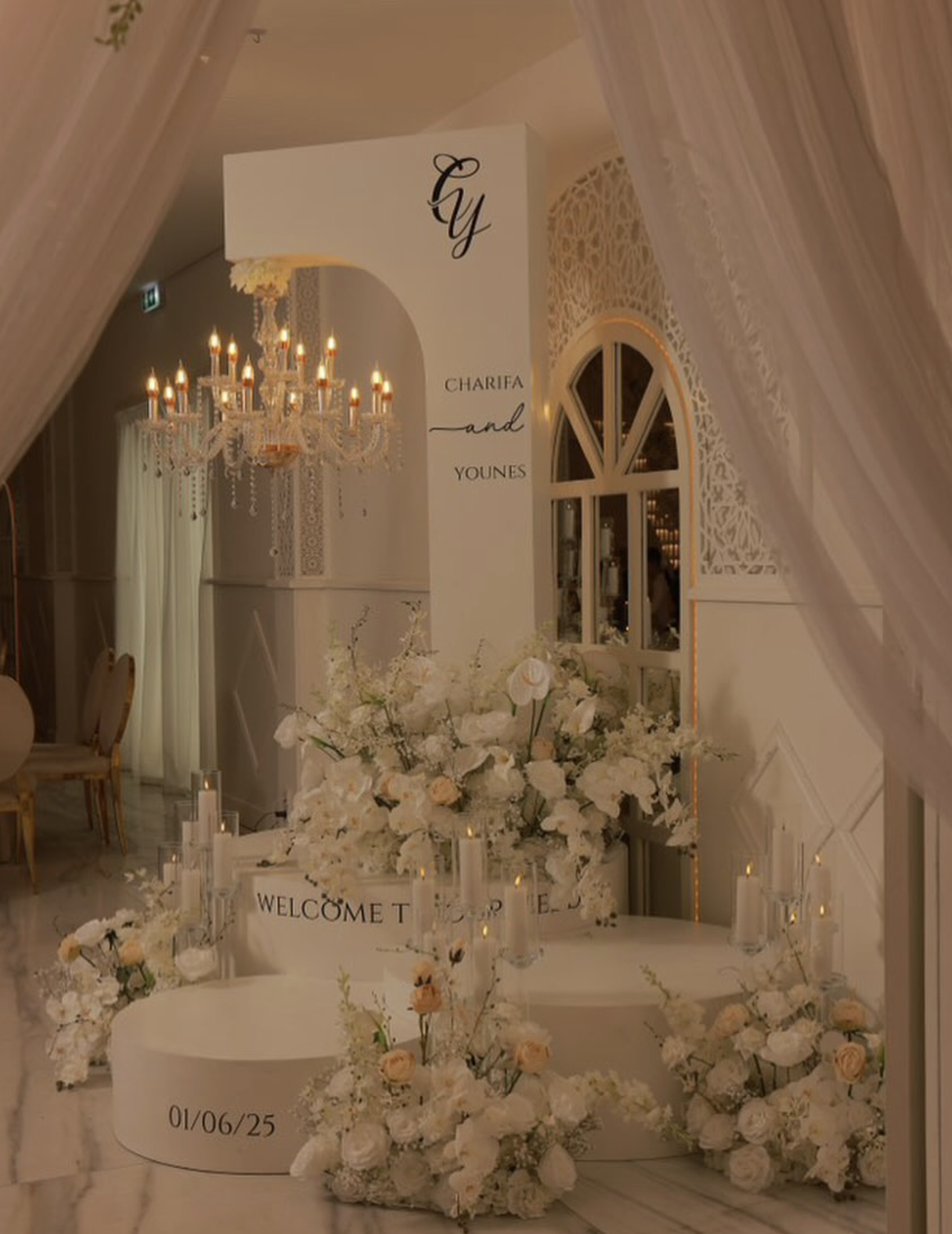 Elegante romantische bruiloft decor met witte bloemen, kaarsen en kroonluchter in een witte ruimte, met tekstborden en de namen Charifa en Younes.
