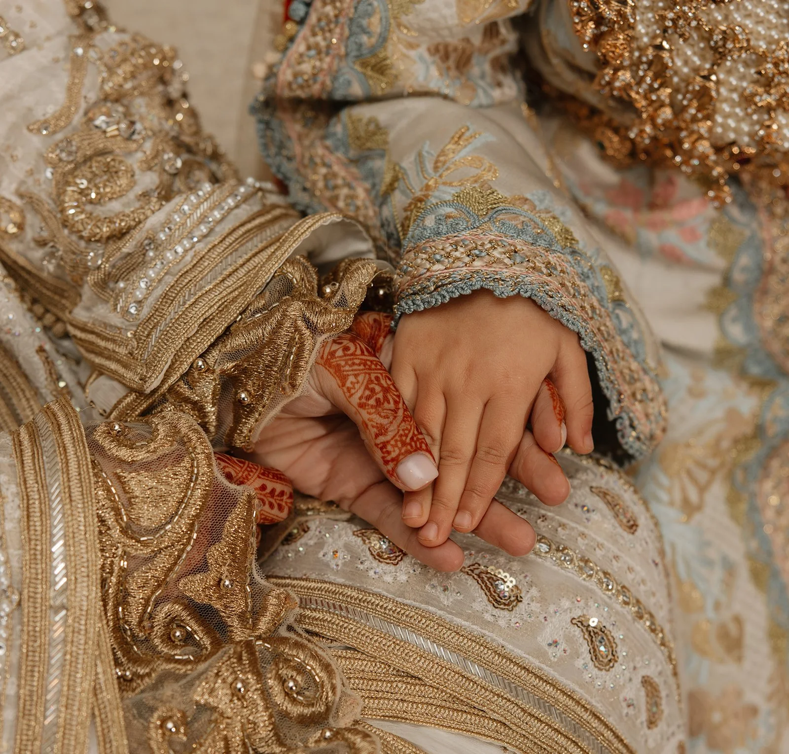 Een close-up van een hand van een volwassene die een jonge hand vasthoudt, beiden in traditionele kleding met ingewikkelde borduursels en glanzende details.
