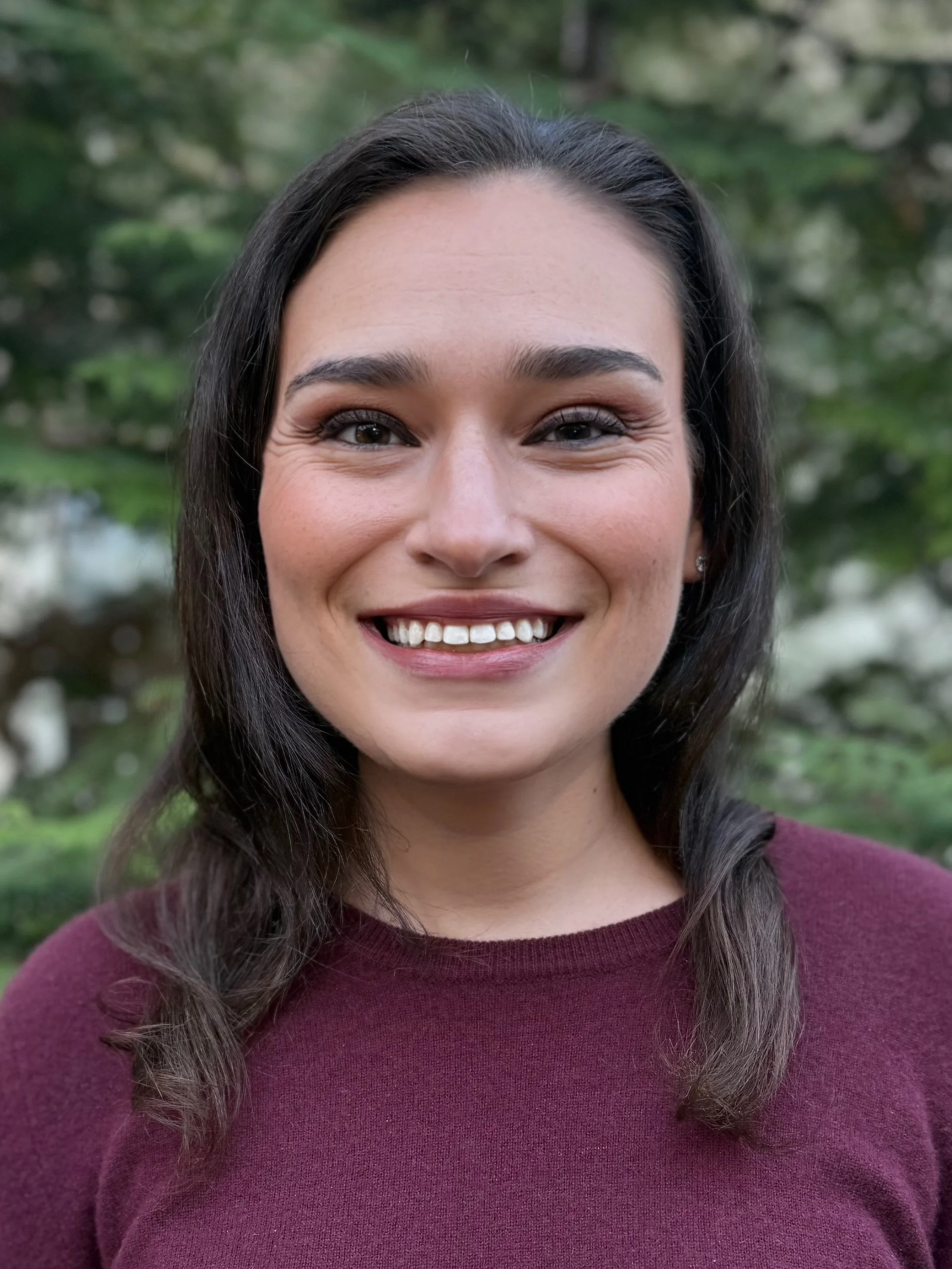 Close-up of a young woman smiling, outdoors with greenery in the background.