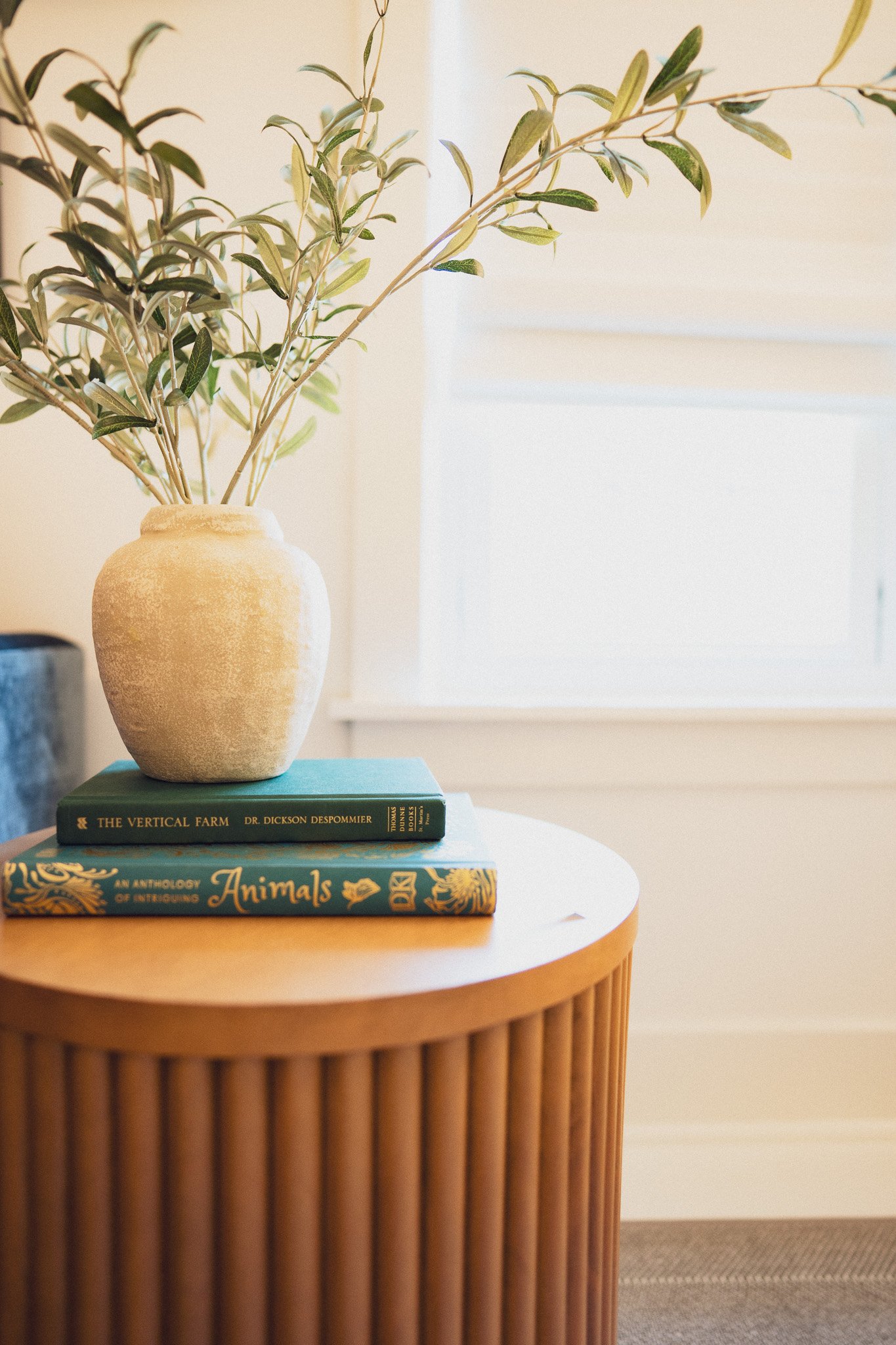 A beige vase with green leaves sitting on top of two books, placed on a round wooden table with vertical ridges.