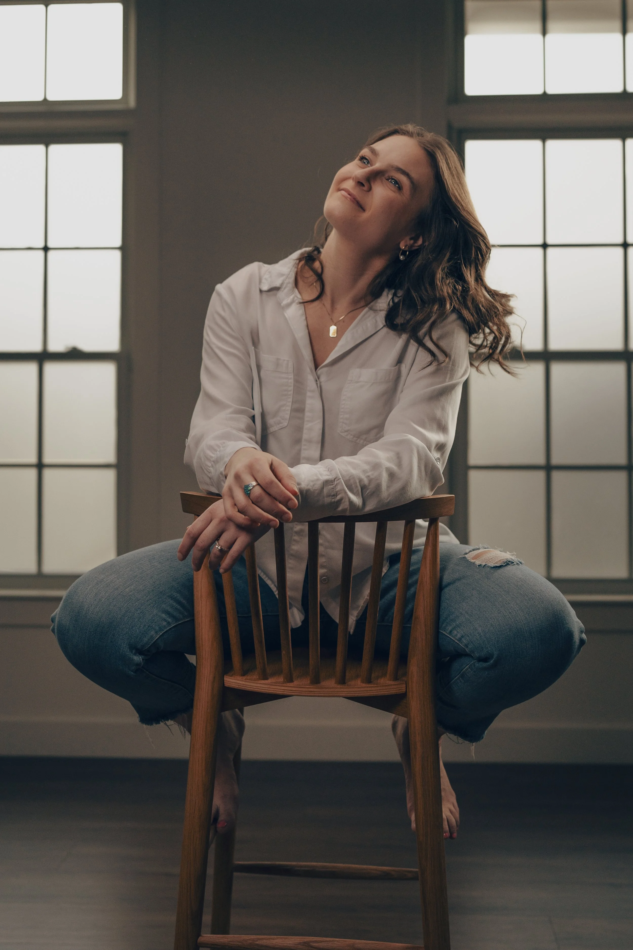 A woman with wavy brown hair smiling and sitting on a wooden chair in front of large windows, wearing a white shirt and ripped jeans.