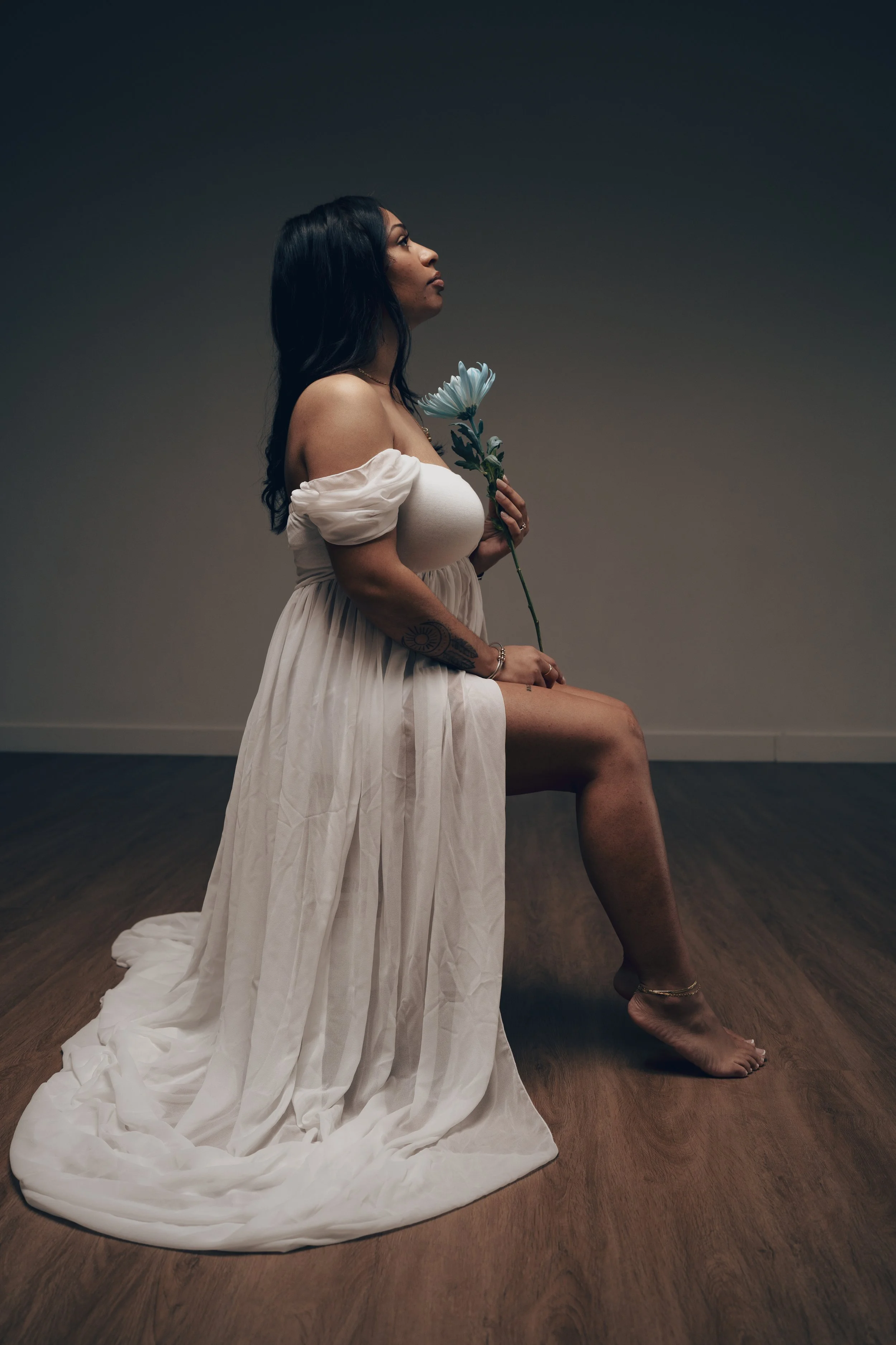 A woman in an off-the-shoulder white dress sitting on a wooden floor, holding a white flower, with a plain gray background.