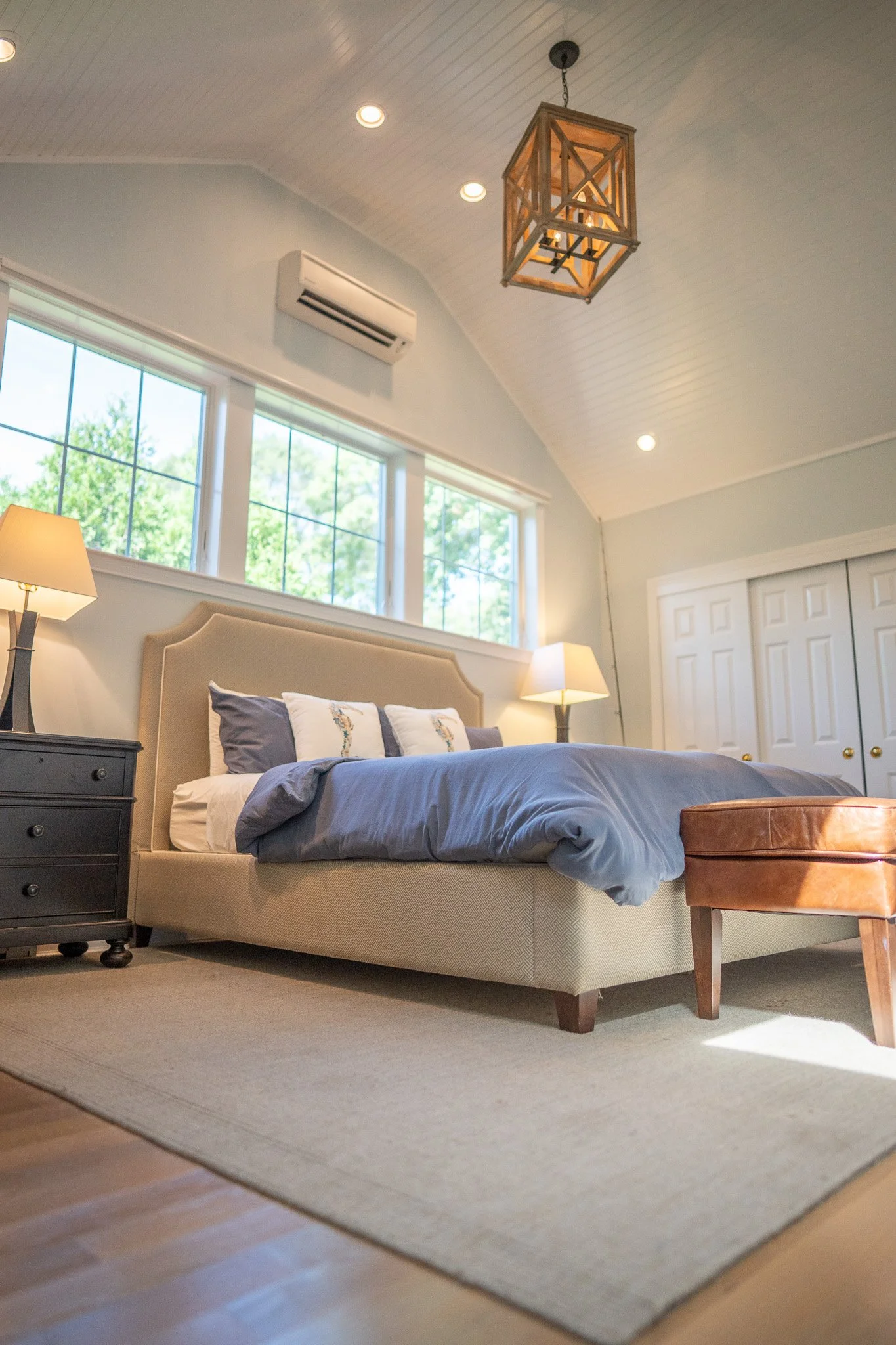 Bright bedroom with large windows, beige upholstered bed with gray and white bedding, two nightstands with lamps, a brown leather ottoman at the foot of the bed, white closet doors, and a wooden ceiling with a geometric pendant light.