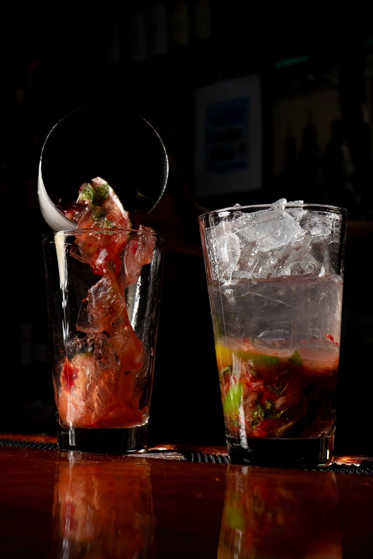 A bartender pours a dark beverage into a glass with ice and fruit, next to another glass filled with ice and fruit.