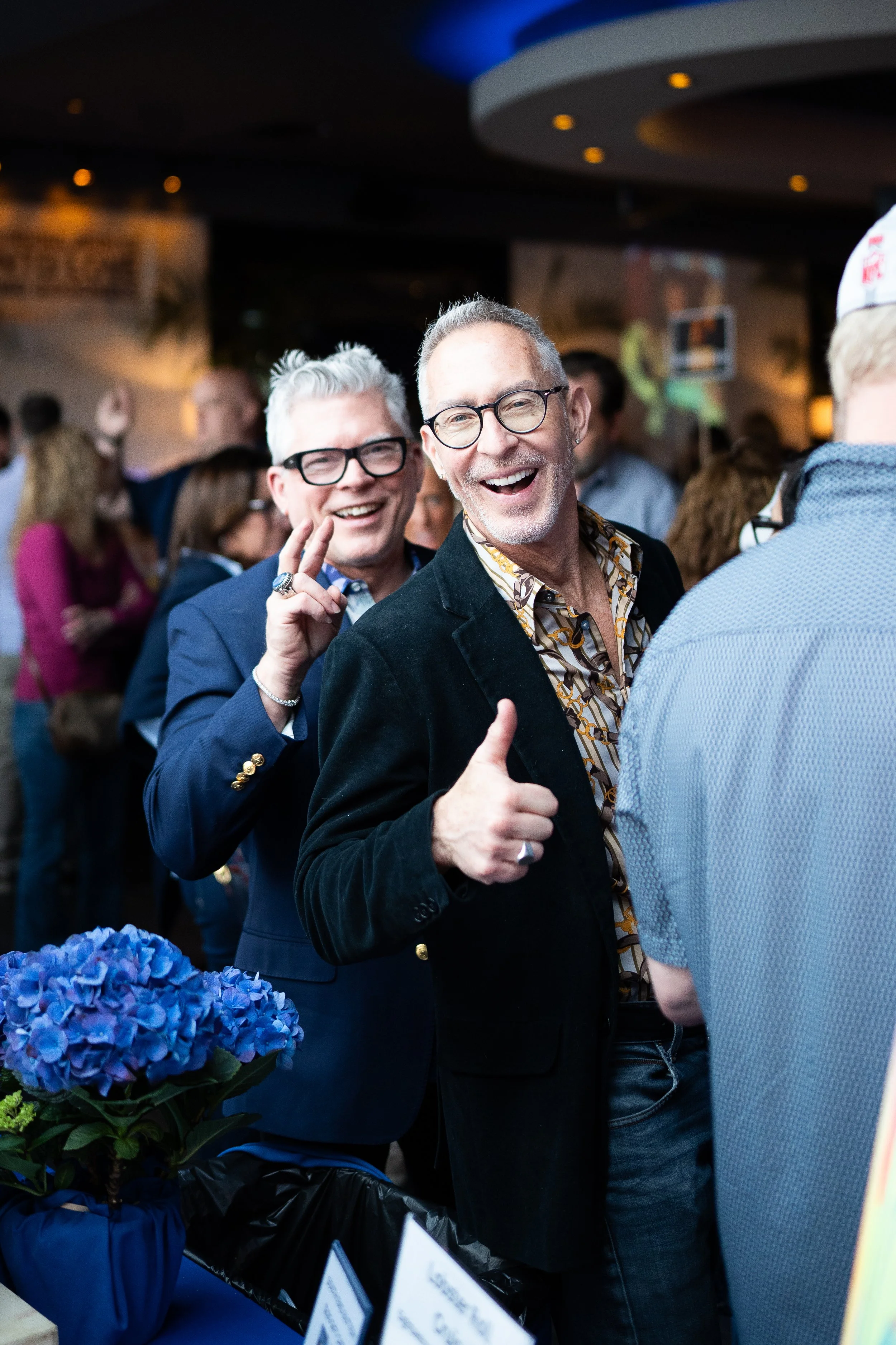 Two men with glasses smiling and giving thumbs up at a crowded event, one with a patterned shirt and blazer, the other with a blue jacket, with blue flowers in the foreground.