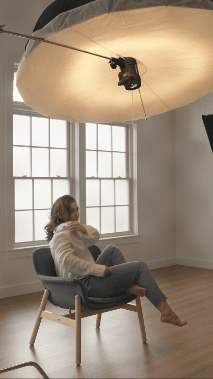 A woman sitting in a modern chair in a bright room with large windows and wooden floors, under a large, studio light diffuser.