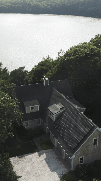 A house with solar panels on the roof, surrounded by green trees, near a large body of water.
