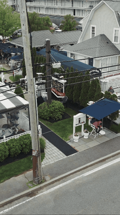 A street view showing a small house with a blue canopy, outdoor seating, and a sign for a bakery or café. There are power lines and trees, with a sidewalk and parked cars in the background.
