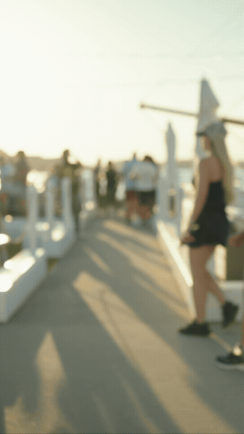 People walking on a dock or pier by the water during sunset, with sailboats and a windmill in the background.