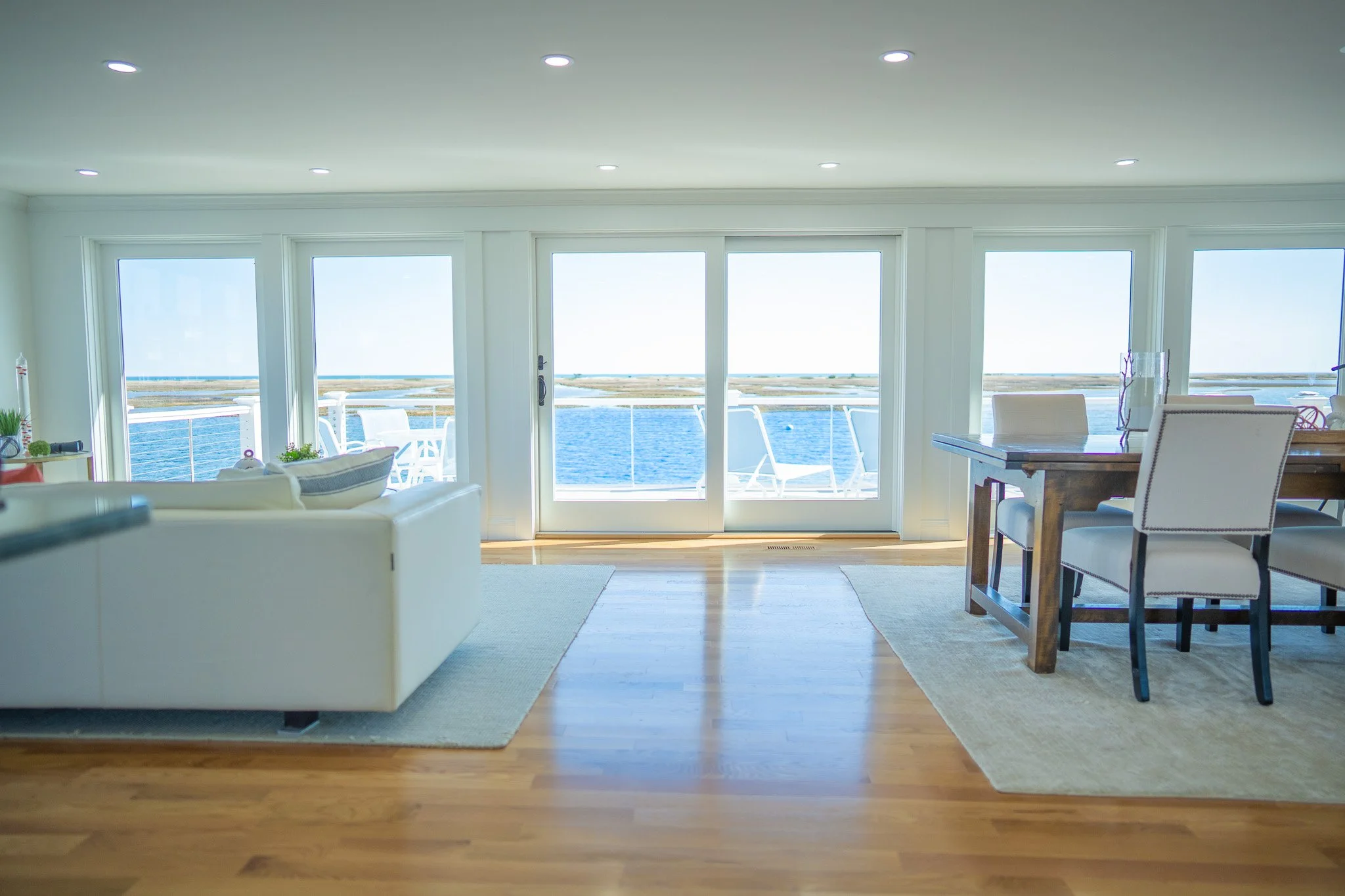 Living room with white sofa, dining table with chairs, large sliding glass doors leading to a balcony with outdoor chairs, overlooking a body of water and distant land.