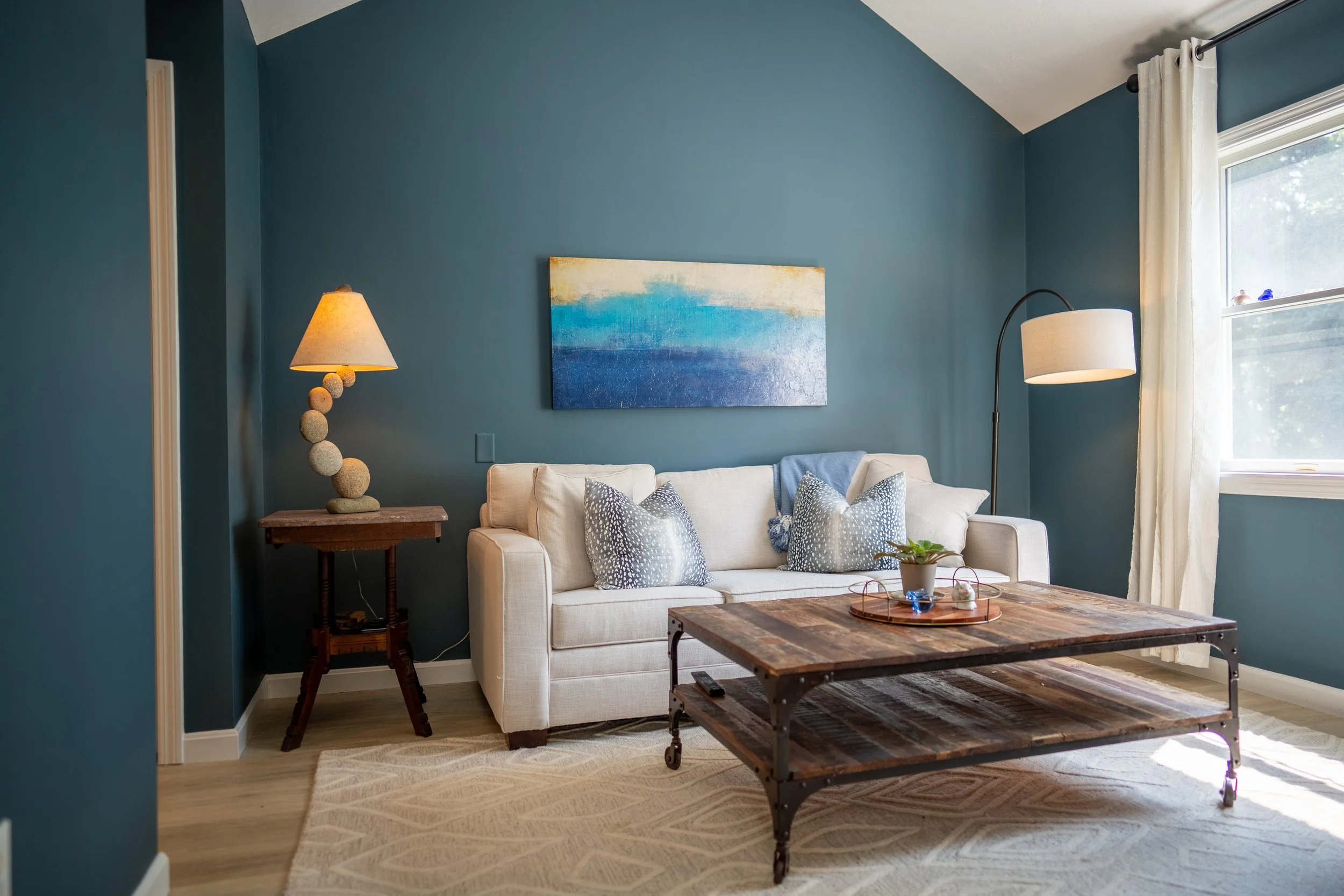 Living room with blue walls, cream sofa with patterned pillows, wooden coffee table, standing lamp, and table lamp near window with white curtains
