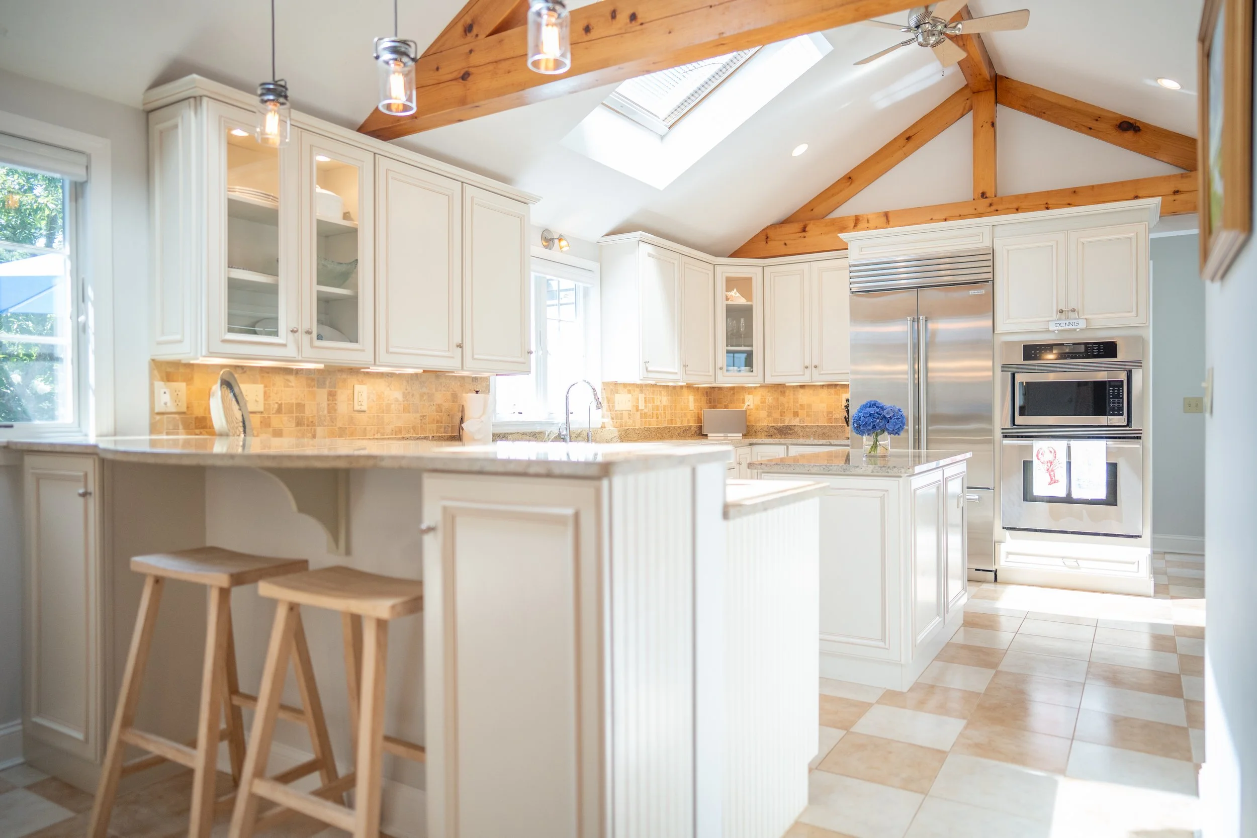 Bright kitchen with white cabinets, a skylight, wooden beams, granite countertops, and stainless steel appliances, including a refrigerator and microwave, with a small island and bar stools.