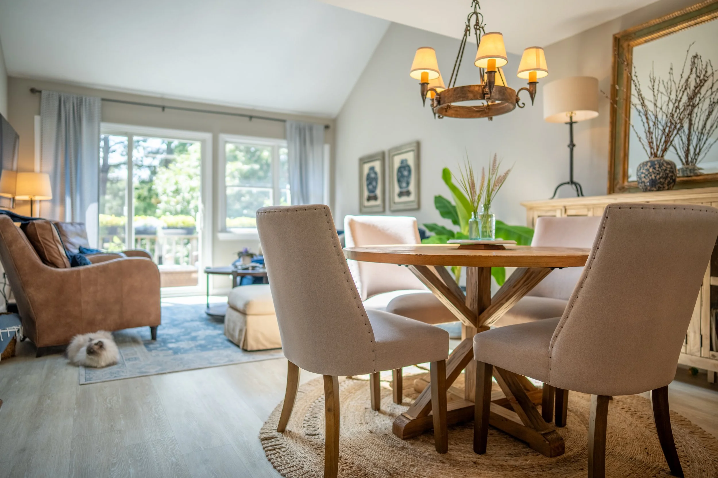 Living room with a wooden dining table and four upholstered chairs, a sofa, a chandelier, a sliding glass door with curtains, and natural light.