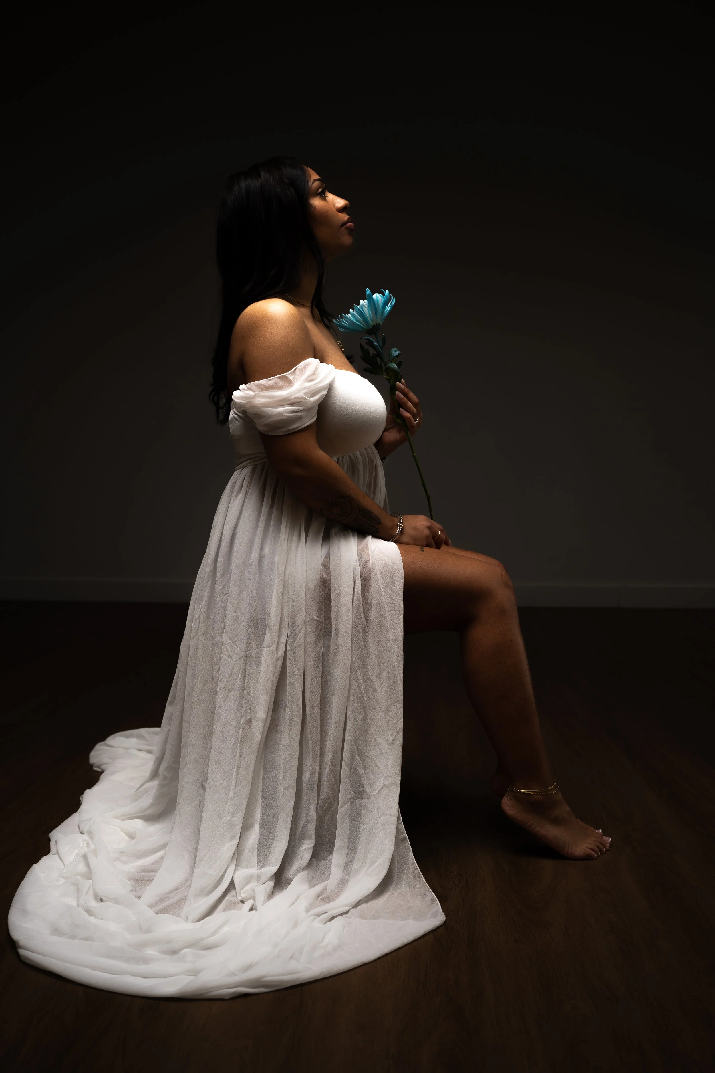 A woman sitting on the ground, wearing a flowing white dress, holding a blue flower, with a dark background.
