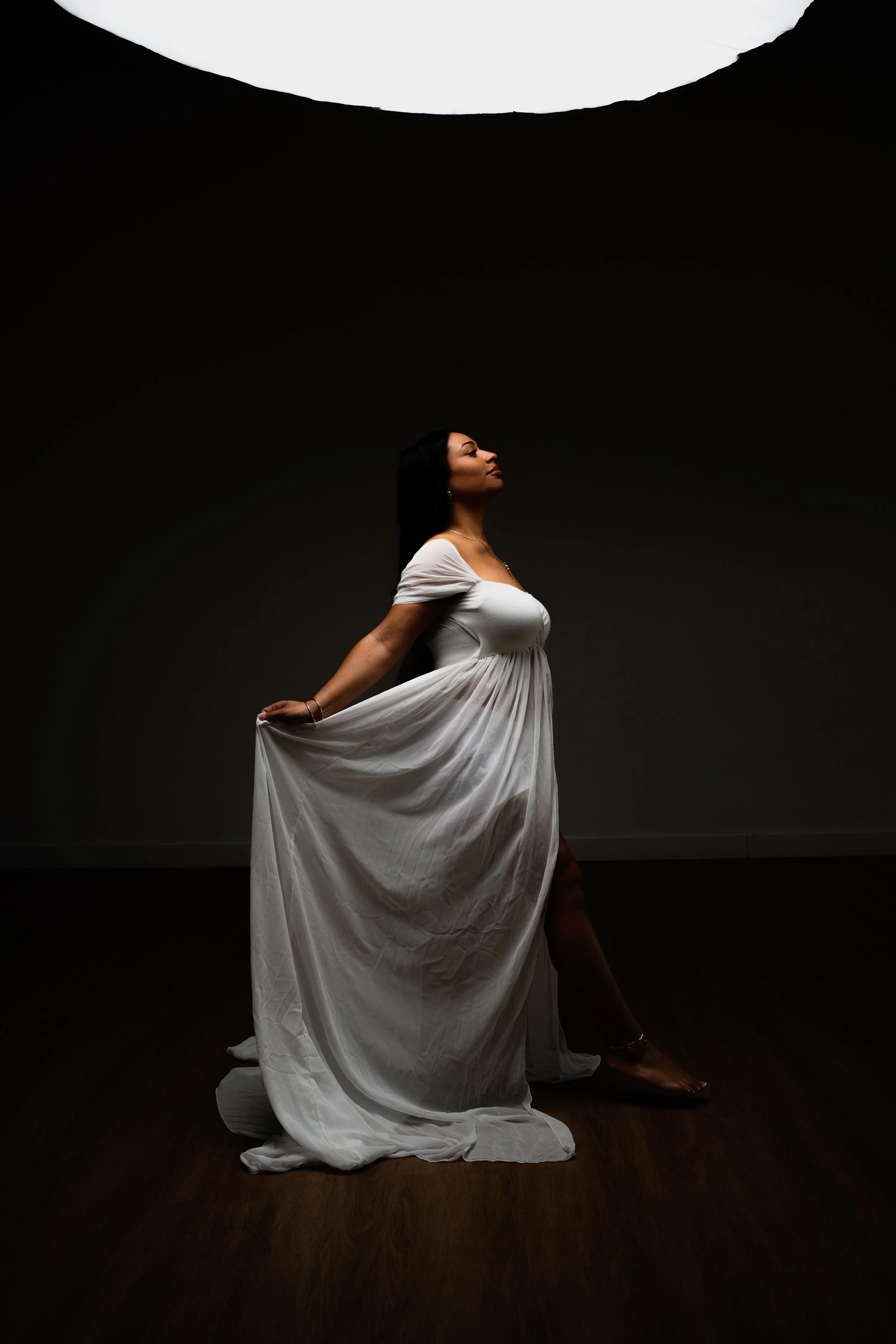 A woman in a flowing white dress standing on a wooden floor with a dark background and a bright light overhead.