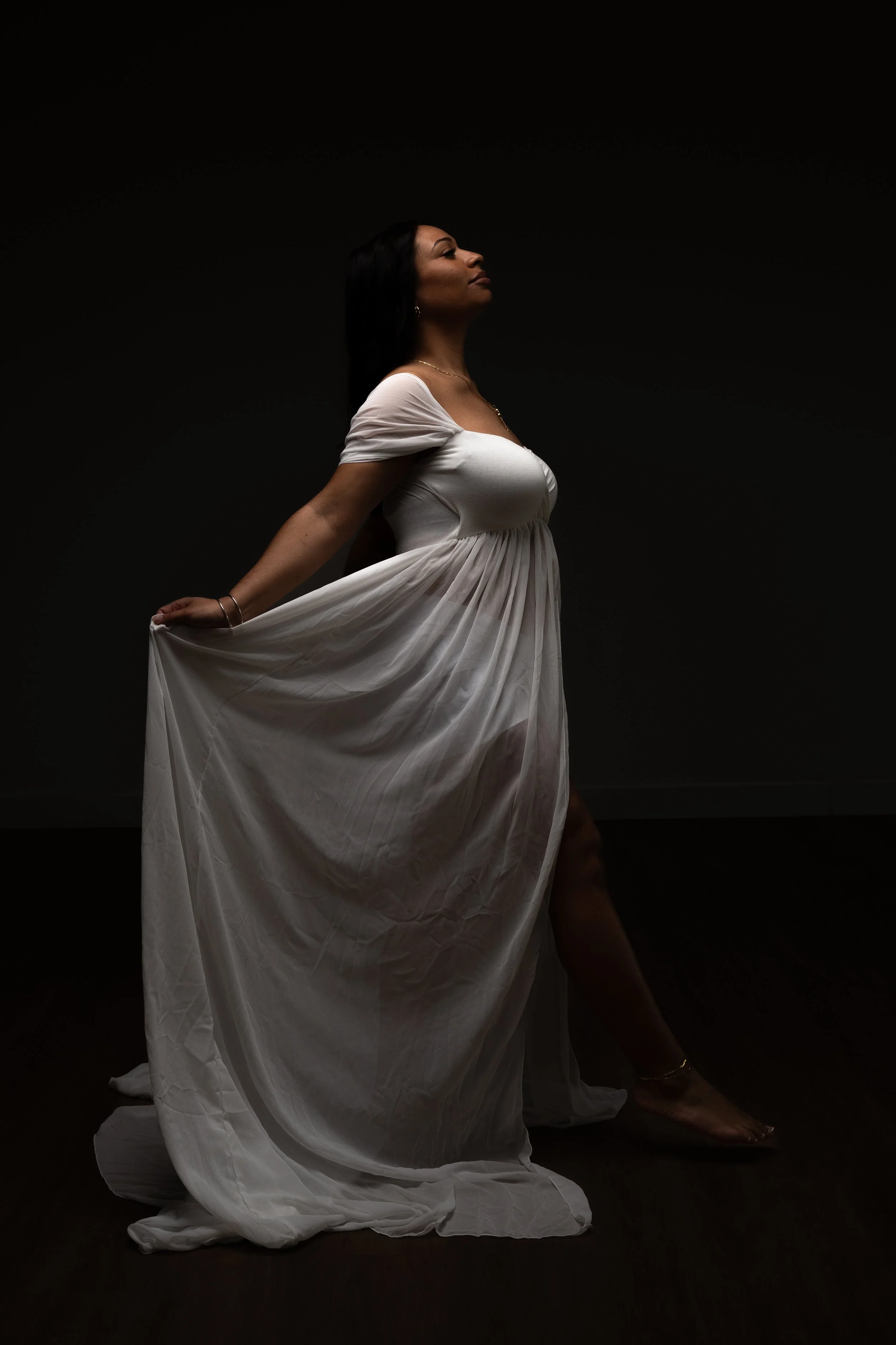 A woman in a white dress poses with one leg bent and her head slightly tilted back against a dark background.