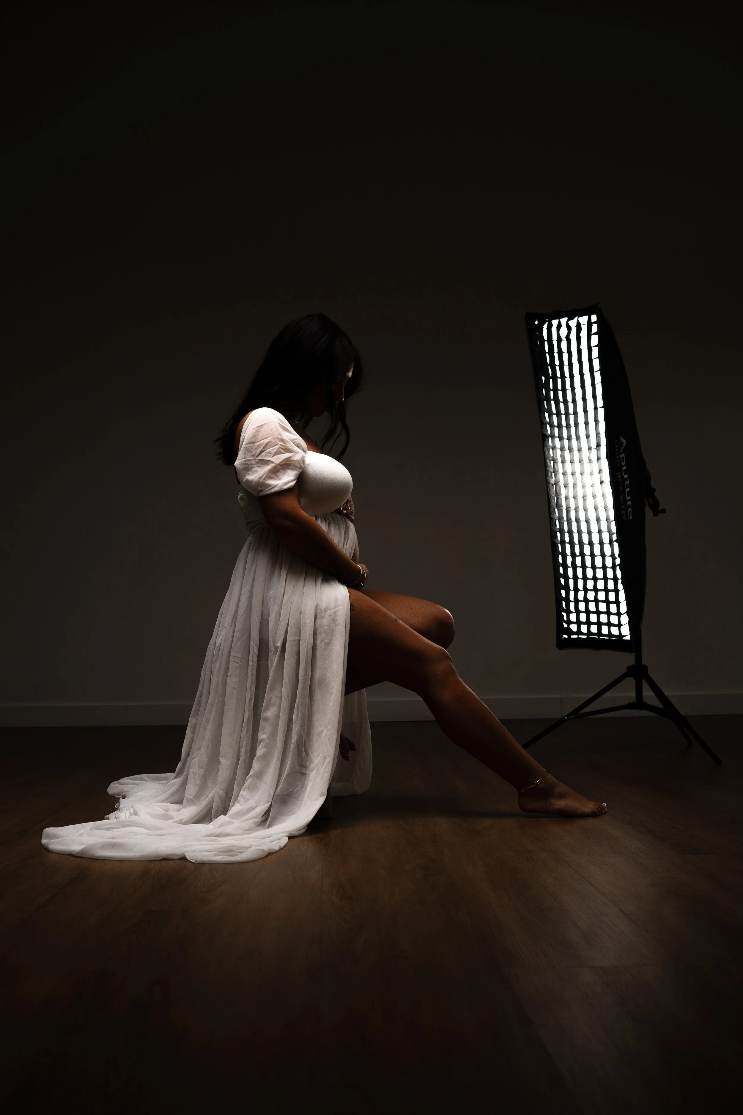 A pregnant woman sitting on a wooden floor in front of a studio light, wearing a flowing white dress.