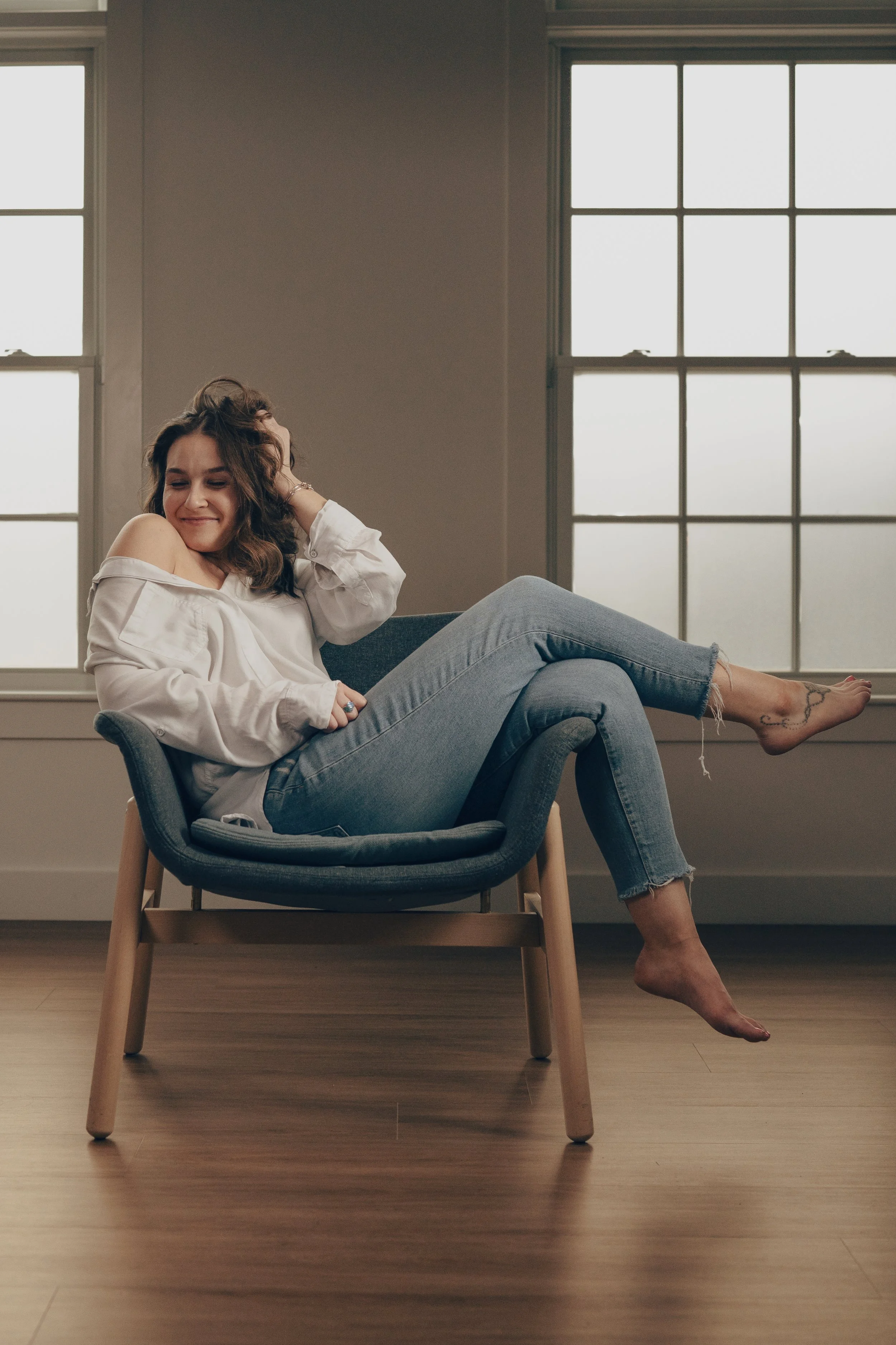 A woman sitting comfortably in a modern chair with wooden legs, smiling and touching her curly hair, in a room with large windows and wooden flooring.