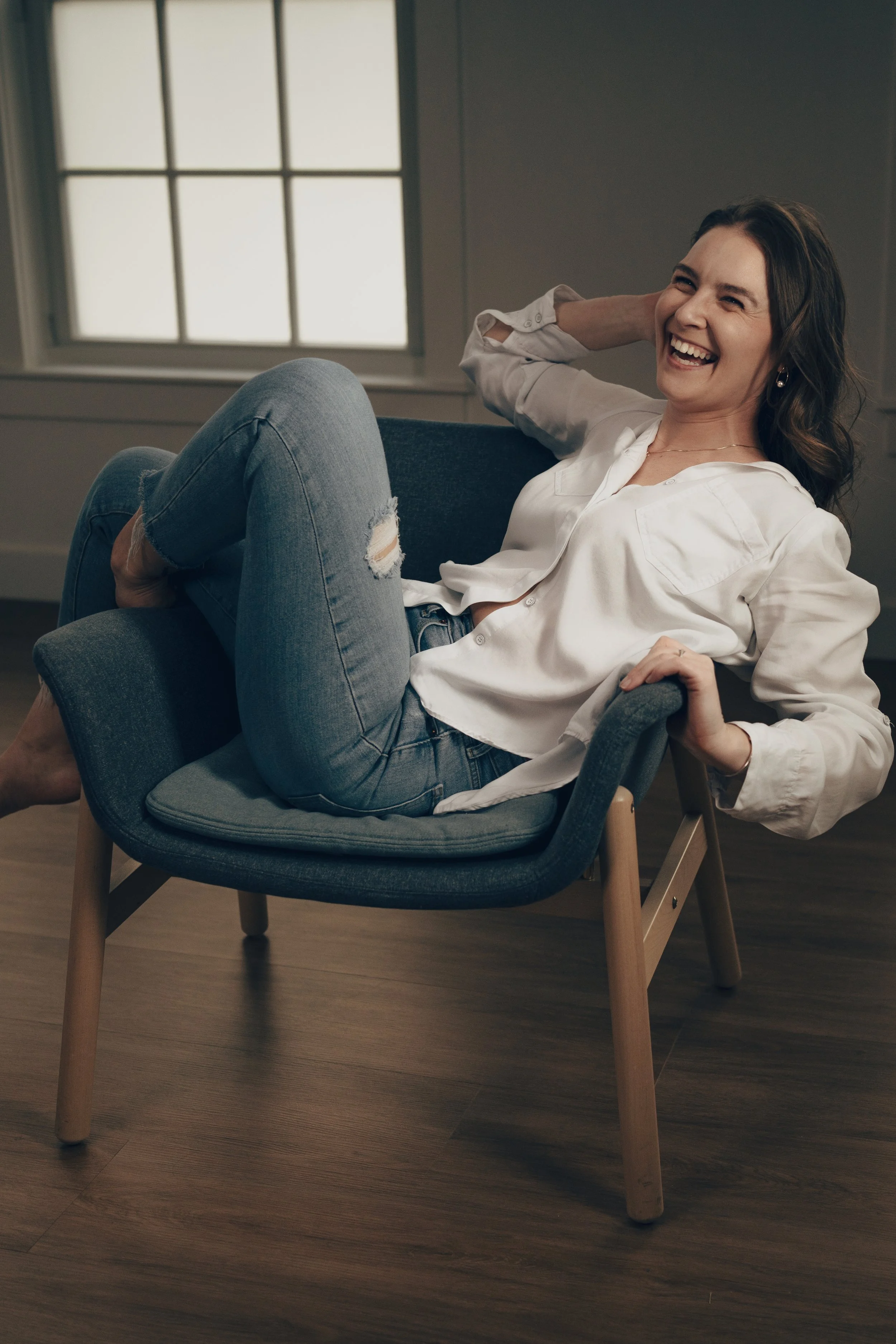 A woman sitting on a modern gray chair with wooden legs, laughing and relaxing in a room with large window panes.