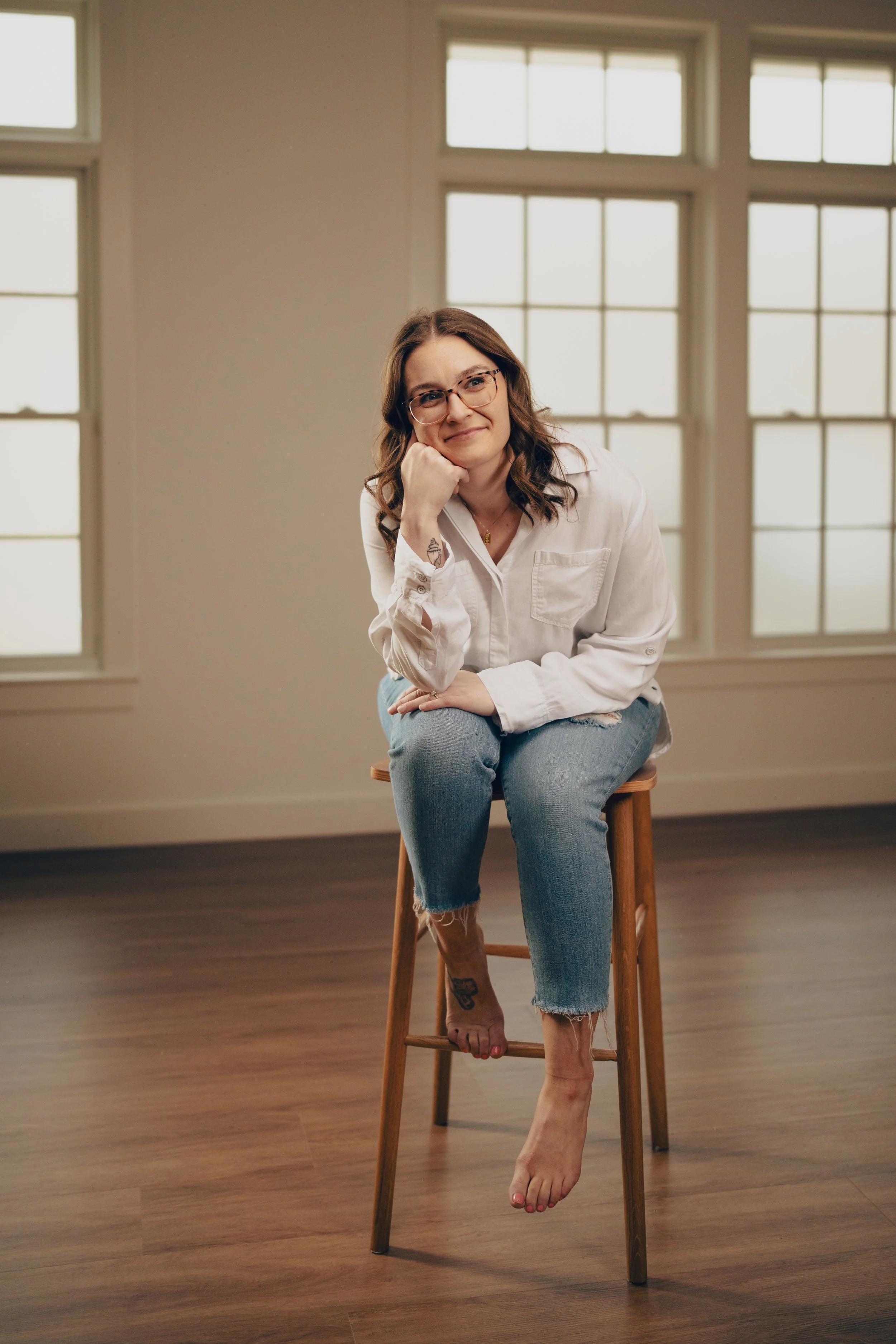 A woman with glasses and shoulder-length brown hair sitting on a wooden stool in a bright room with large windows, wearing a white shirt and ripped jeans.