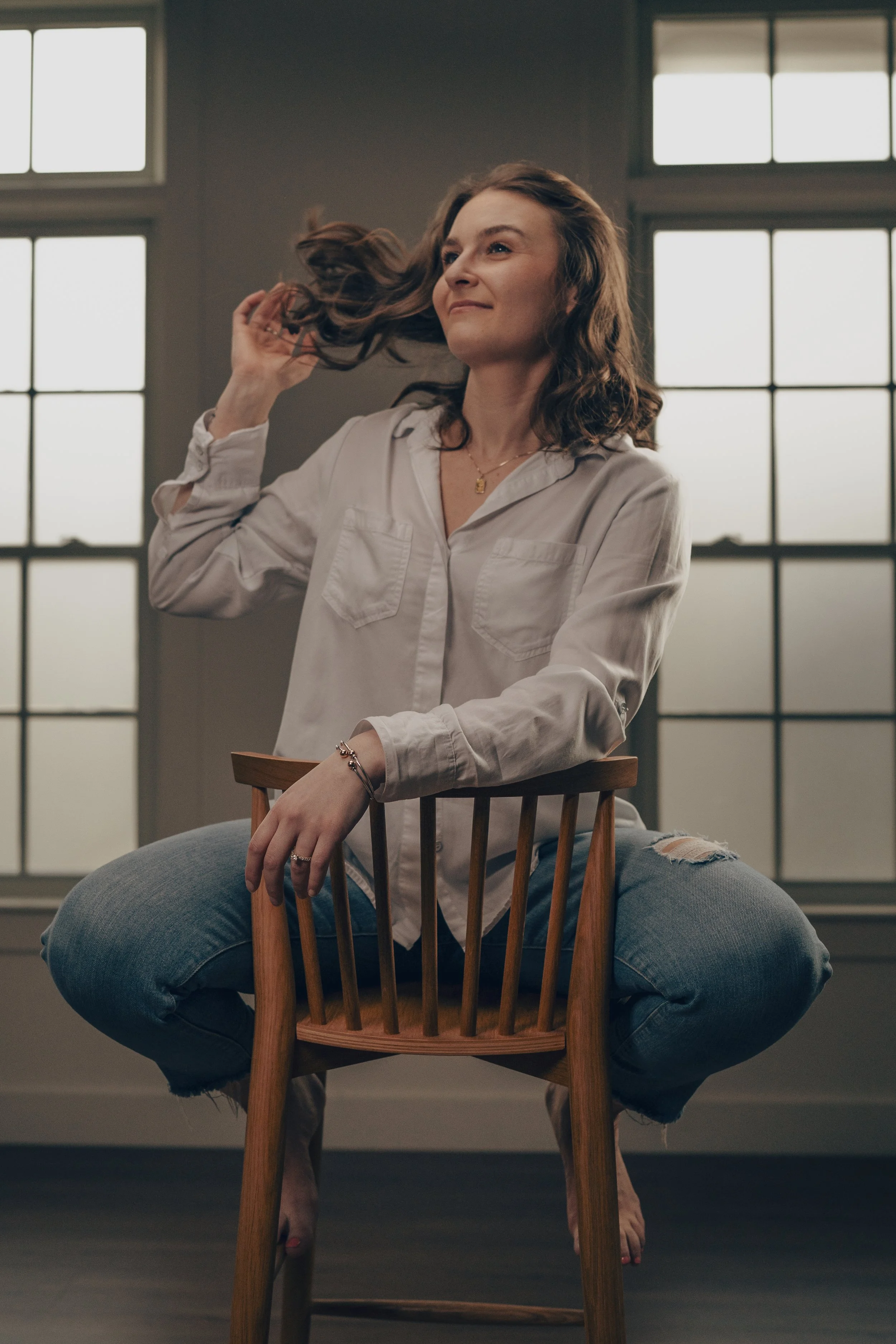 A woman sitting on a wooden chair, lifting her hair with a confident expression, in front of large windows.