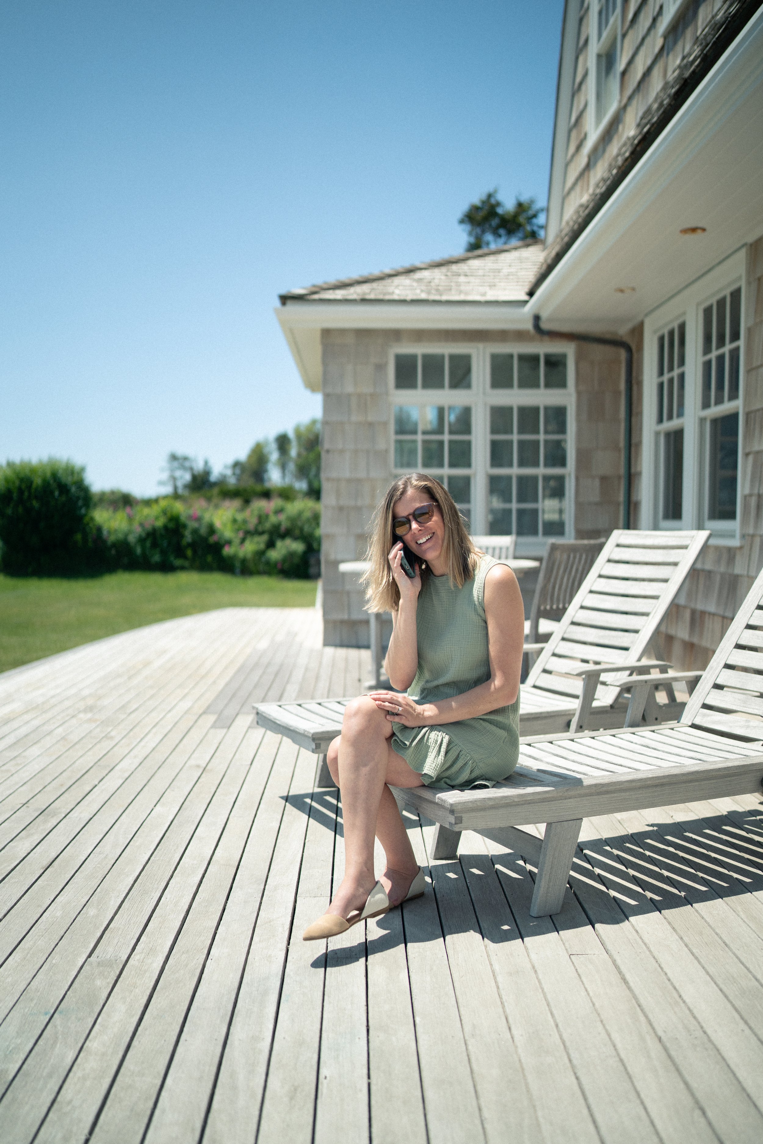 Woman sitting on a wooden deck in front of a house, talking on her cell phone, wearing sunglasses and a green dress, smiling.