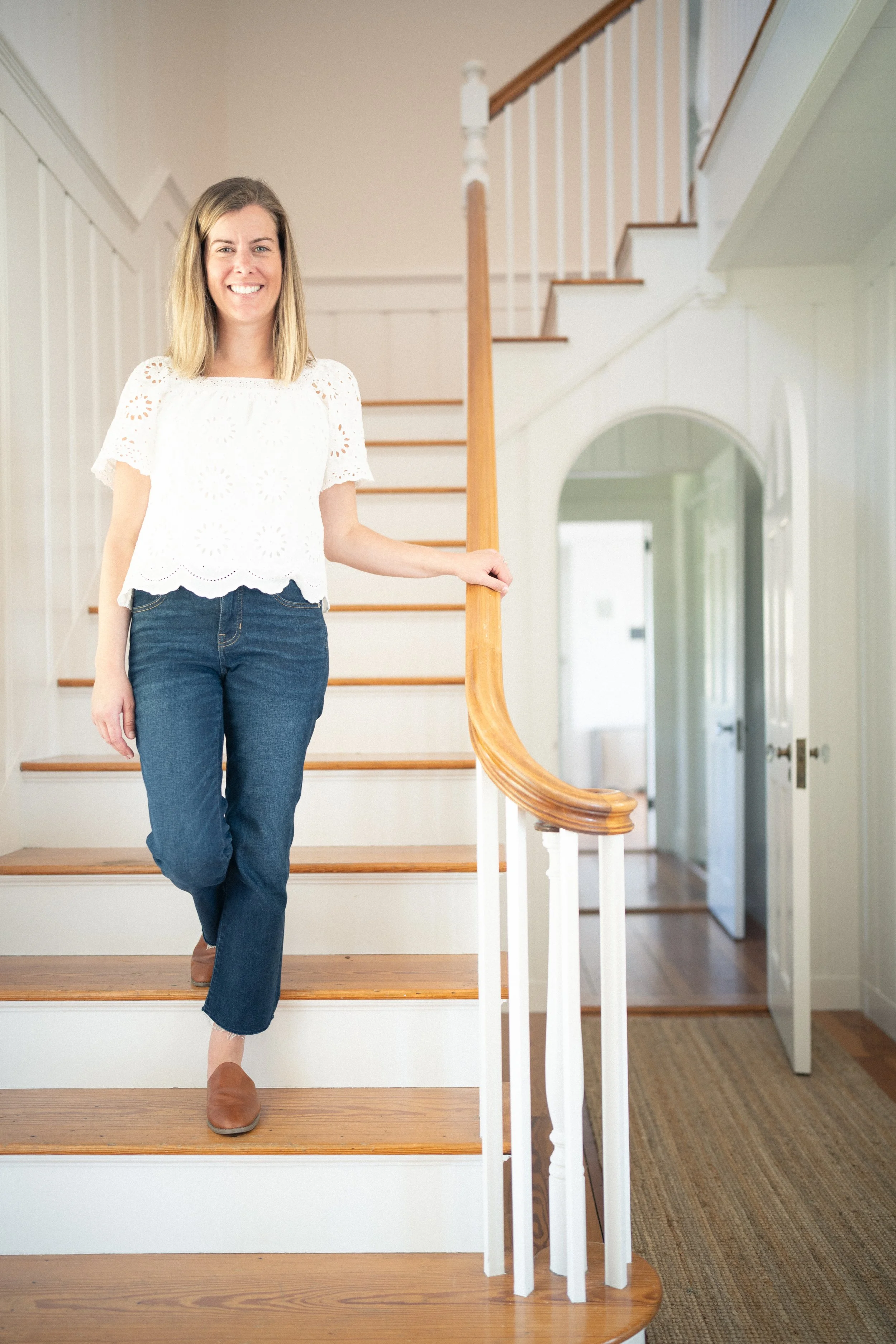 A woman with shoulder-length blonde hair, wearing a white eyelet top and blue jeans, standing on a staircase indoors, holding onto the wooden handrail and smiling at the camera.