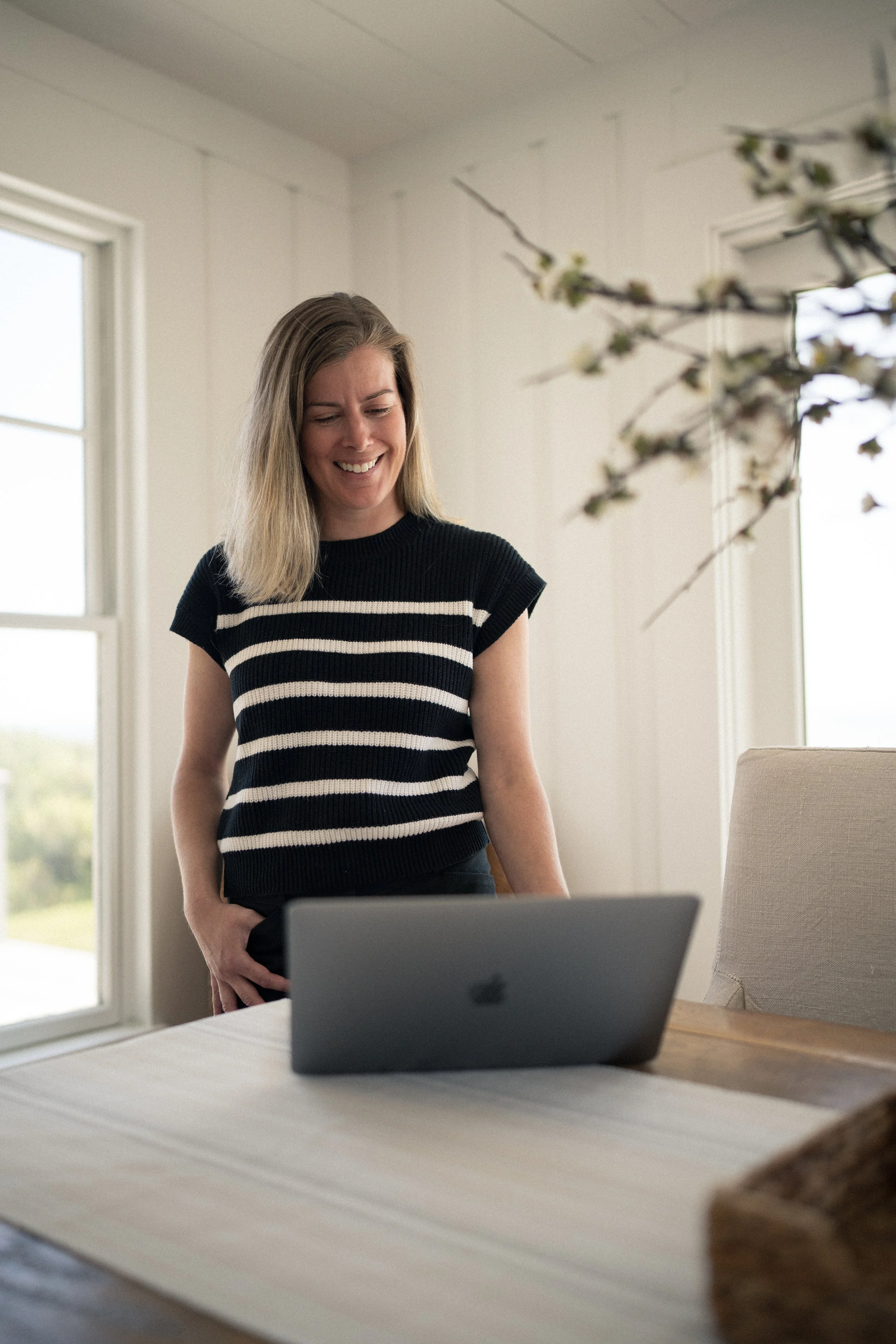 A woman in a black and white striped shirt smiling and looking at a closed laptop on a wooden table in a bright room with white walls and large windows.