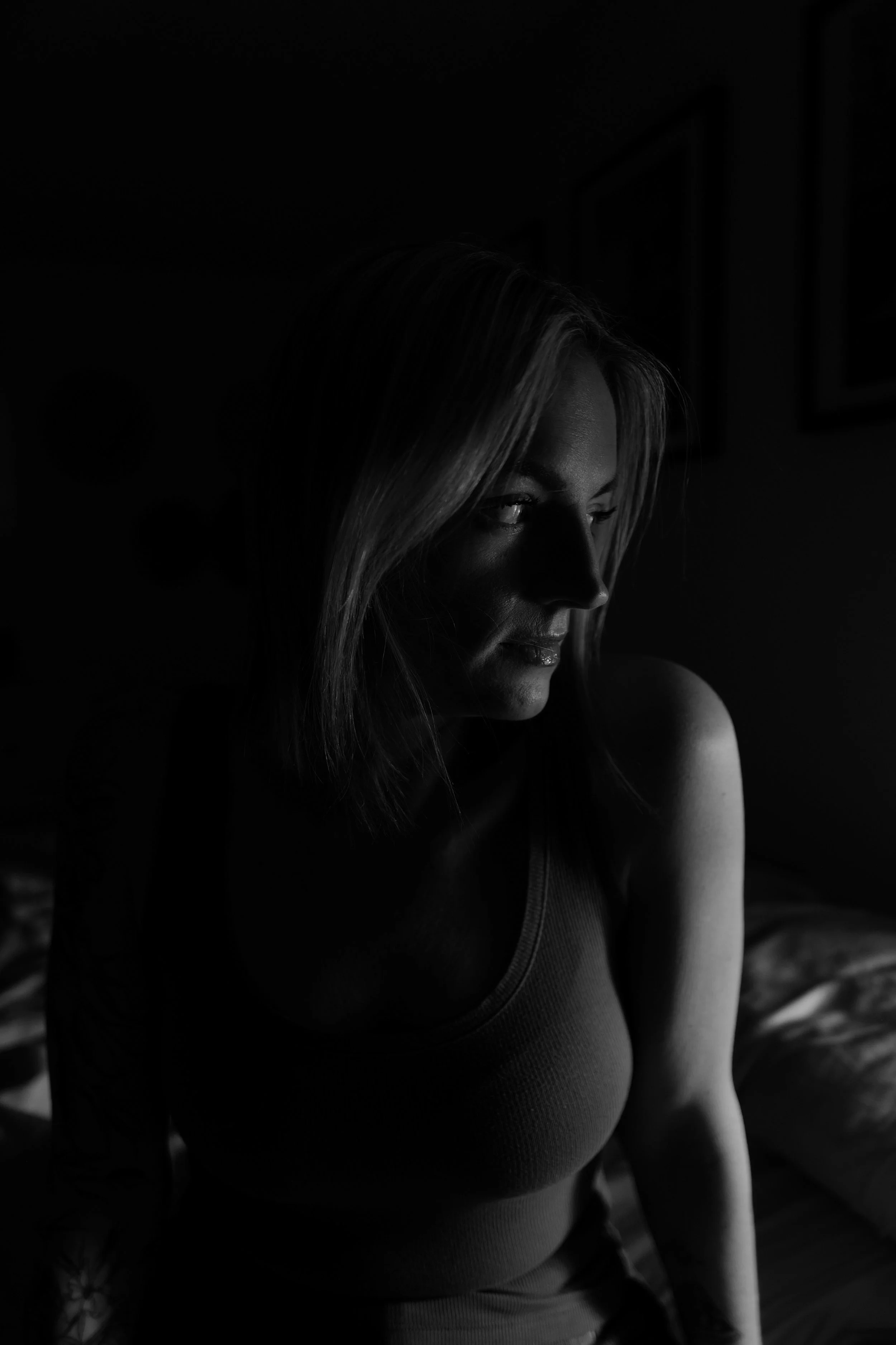 A black and white portrait of a woman with shoulder-length hair, looking down and slightly to the side, in a dimly lit room.