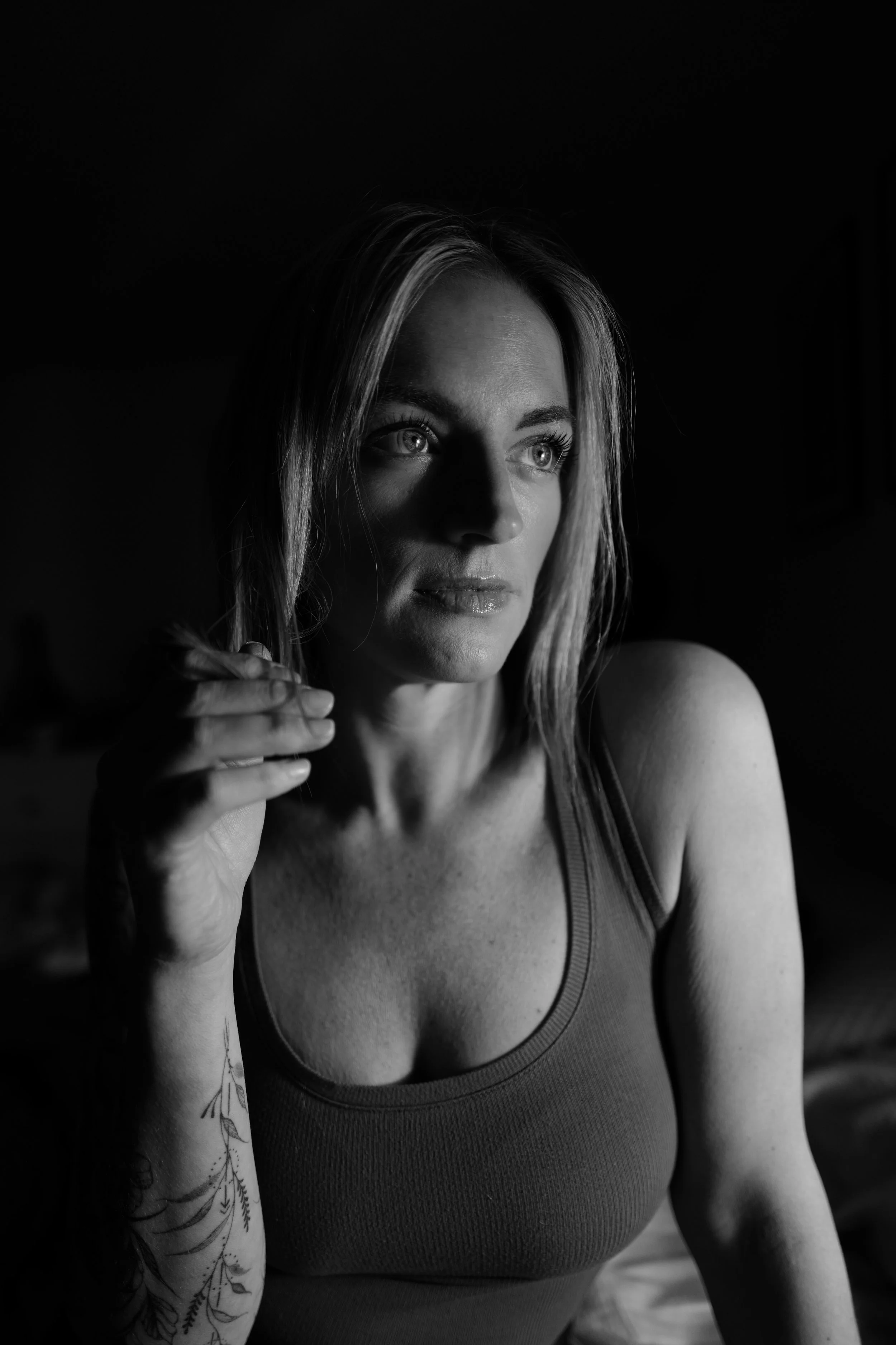 A black and white portrait of a woman with light-colored hair, wearing a sleeveless top, looking pensively to the side with light illuminating her face.