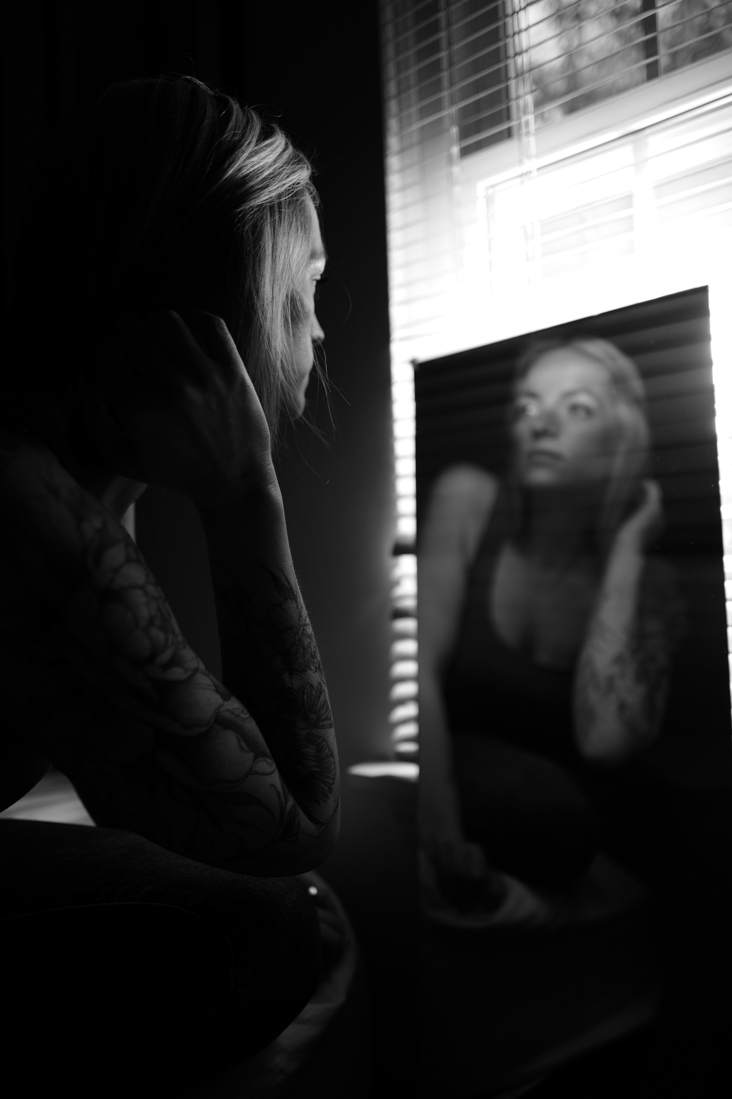 A woman with tattooed arms sitting in a dark room, looking at her reflection in a mirror, with light coming through window blinds.